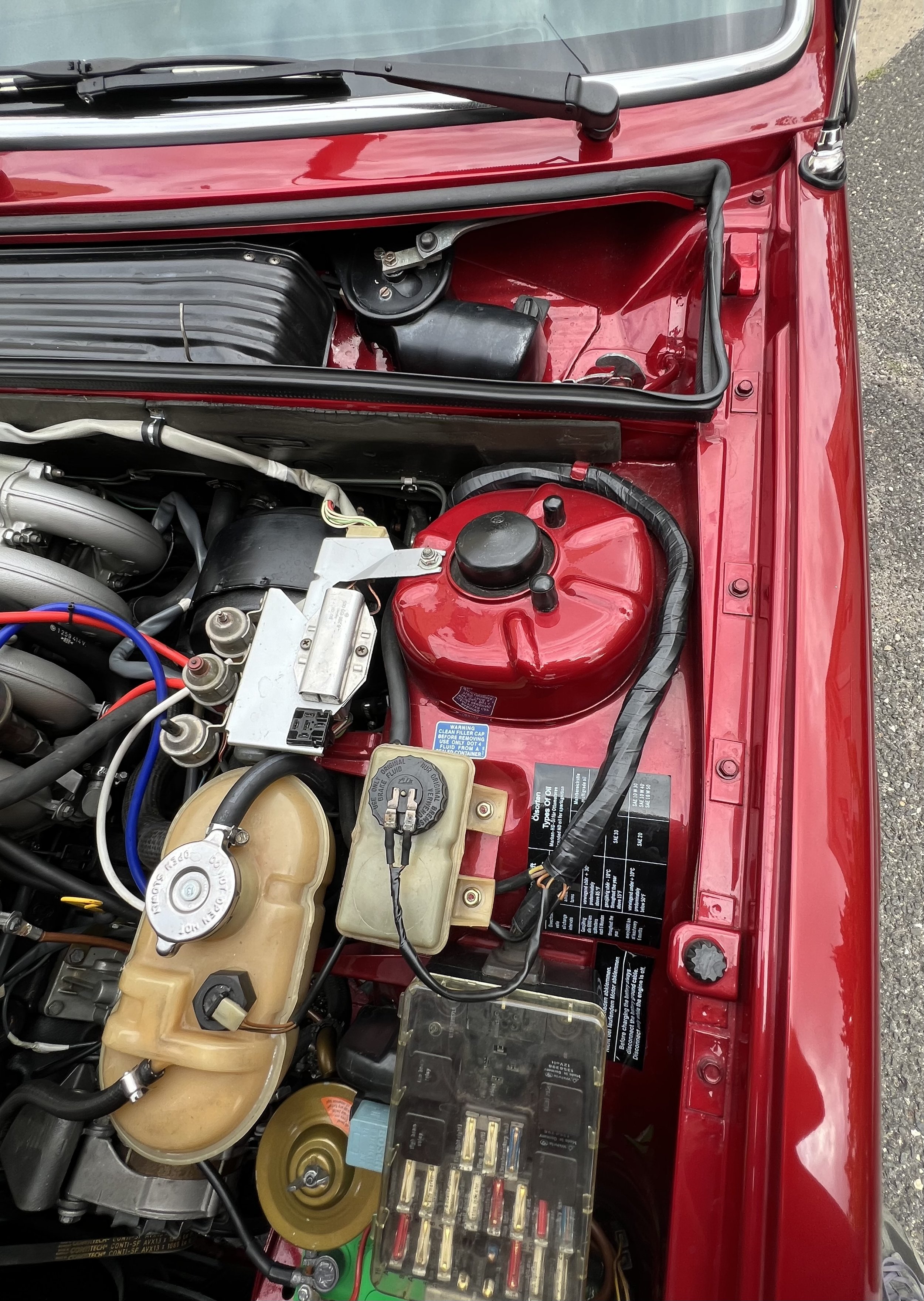 Close-up of a 1977 BMW 630 csi car engine bay with various components, including a red coolant reservoir, fuse box, and other engine parts under the hood.