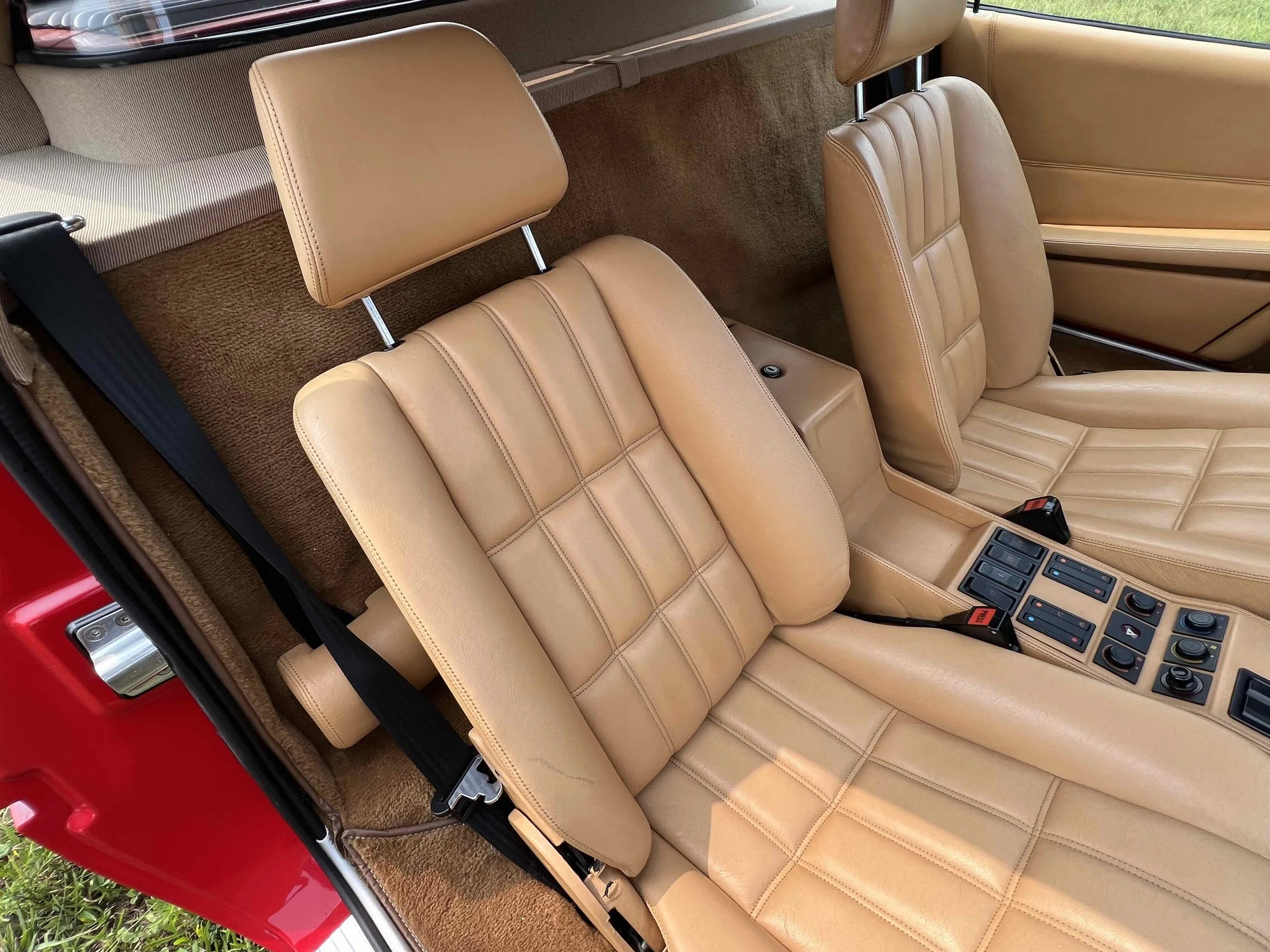 The interior of a vintage sports car with tan leather bucket seats, center console with switches and controls, and black seat belts.