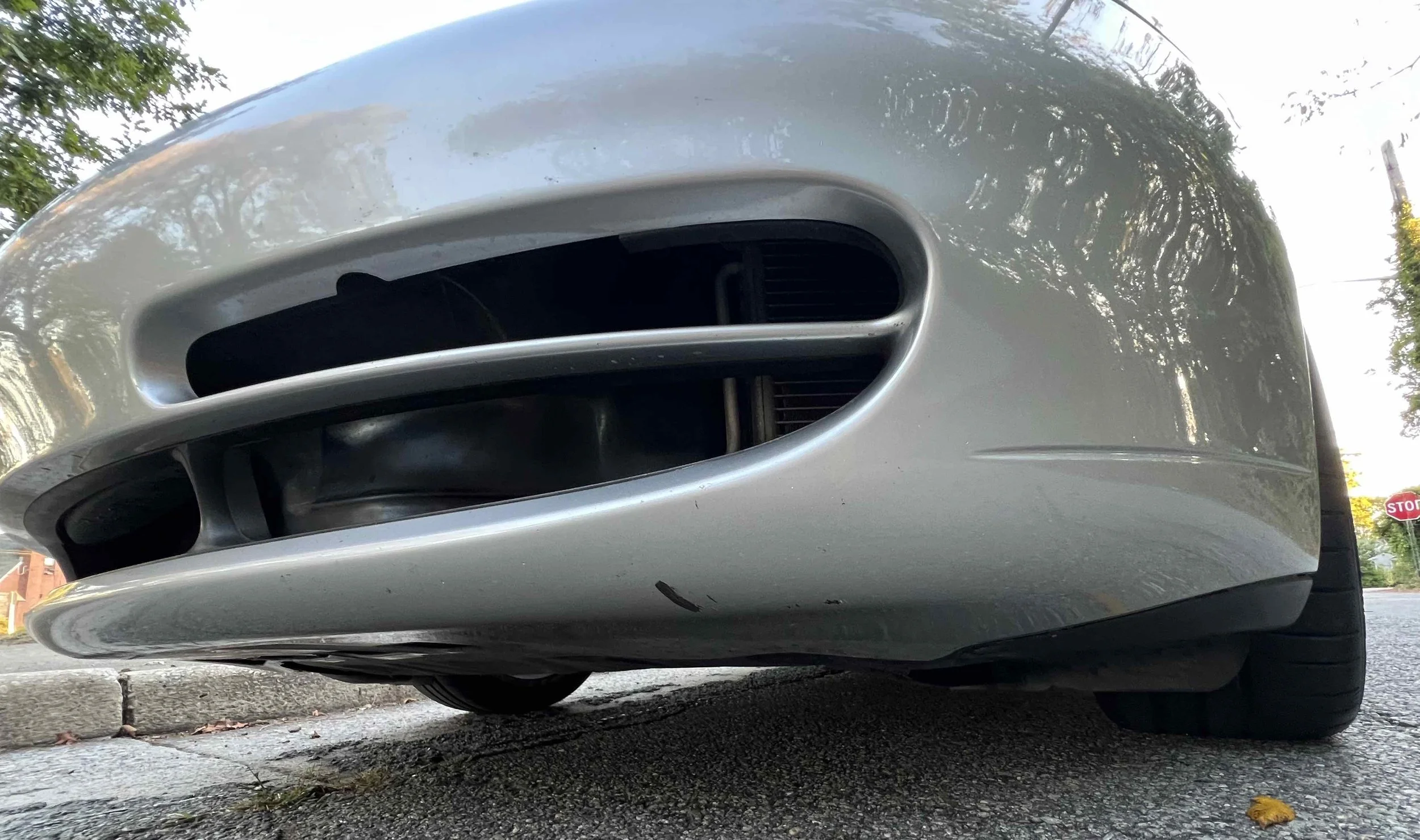 Low-angle close-up of a silver car's front bumper and grille, showing the lower part of the vehicle on an asphalt street with trees and a stop sign in the background.
