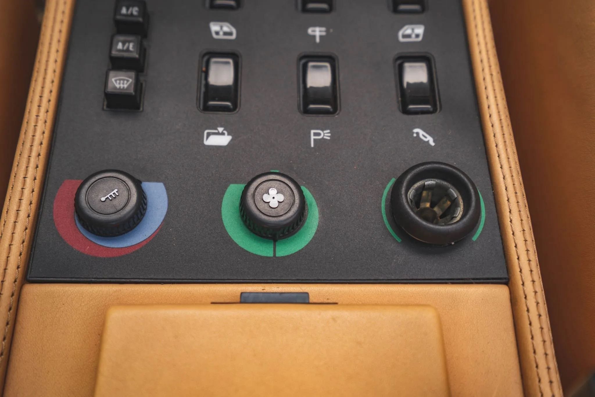 A car's climate control panel with three knobs for temperature, fan speed, and vent direction, surrounded by tan leather trim.
