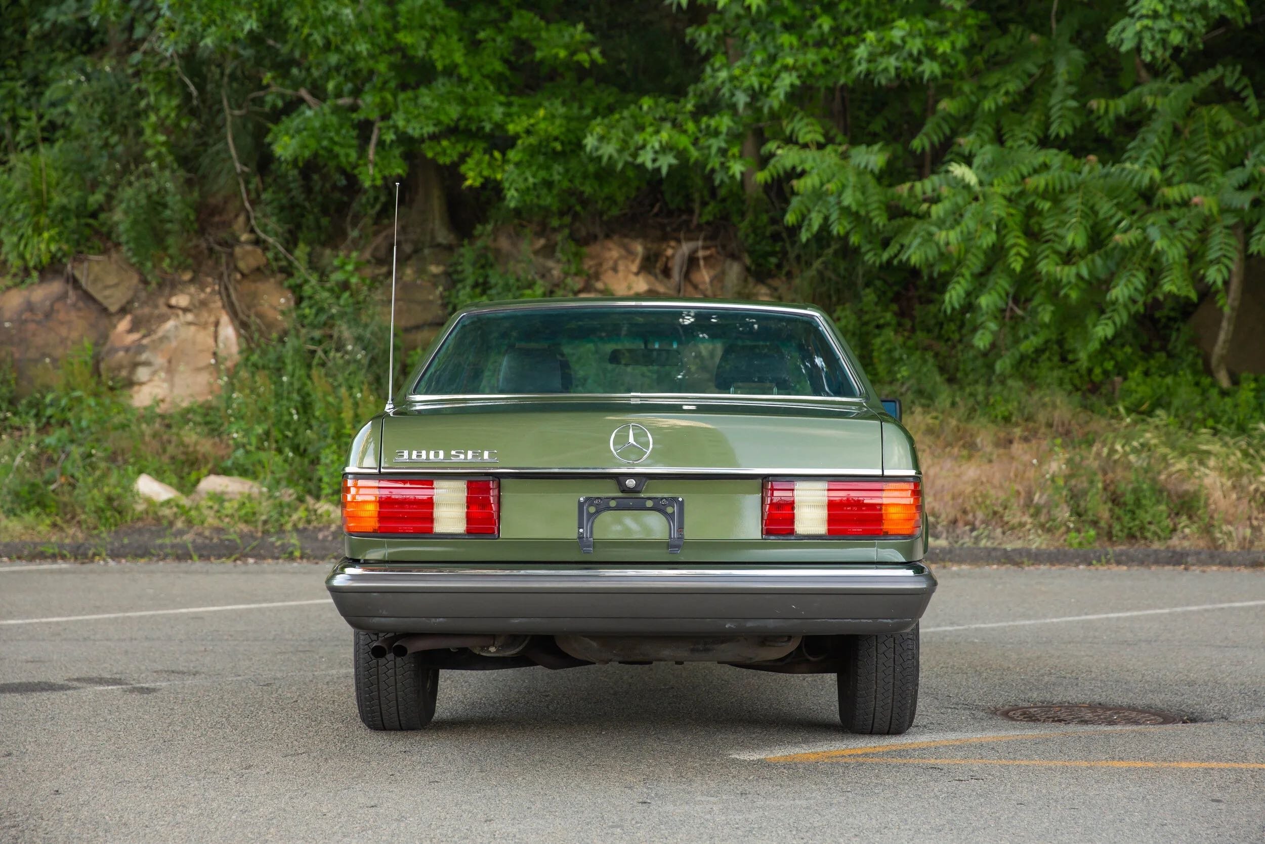 Green Mercedes-Benz 380 SEC car parked on a paved lot with green foliage and rocks in the background.
