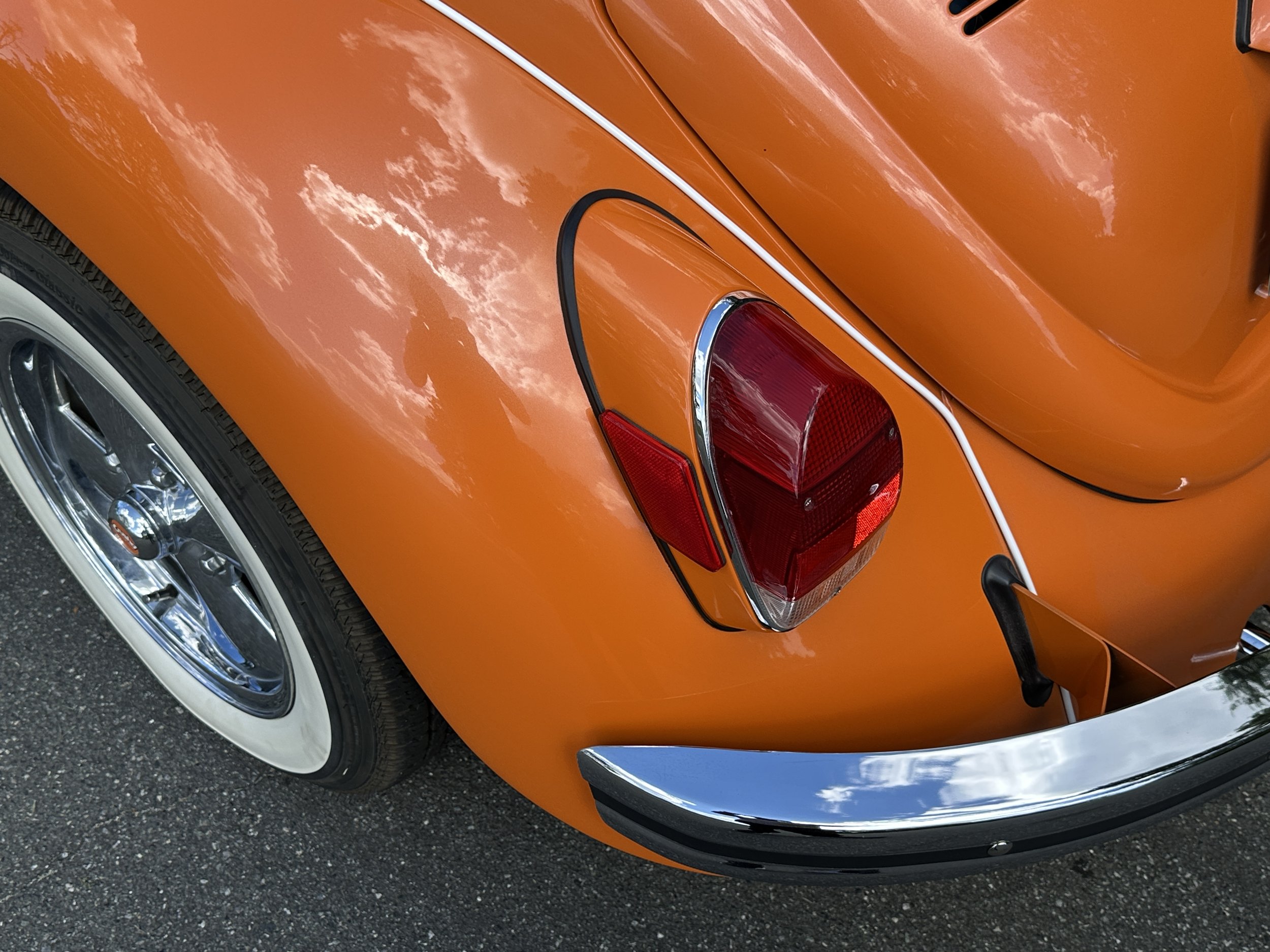 Close-up of the rear side of an orange vintage car, showing a red tail light, chrome bumper, and chrome wheel with a white wall tire.