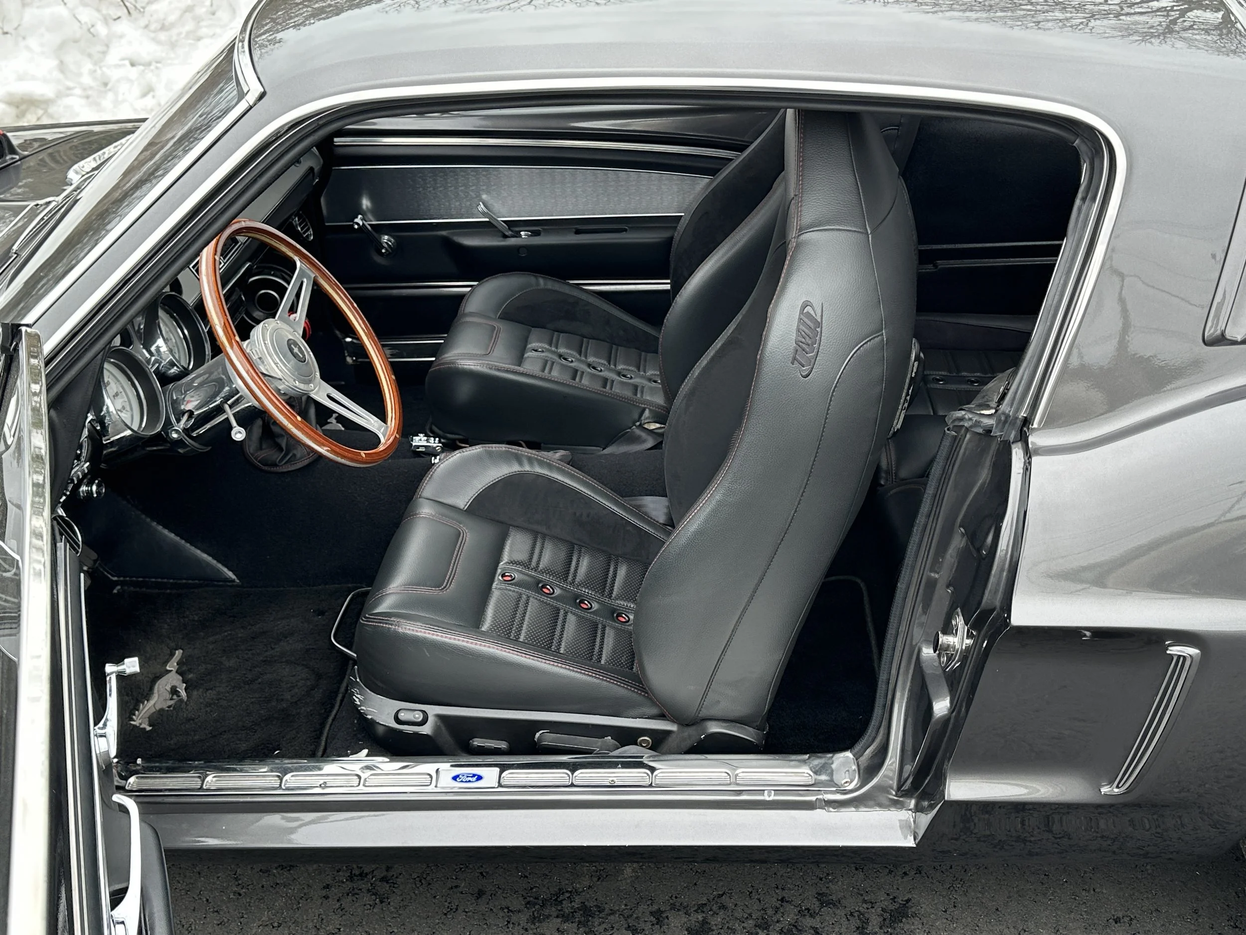 Interior of a vintage Ford race car, featuring black leather seats with red stitching, a wooden steering wheel, and a classic dashboard with analog gauges.