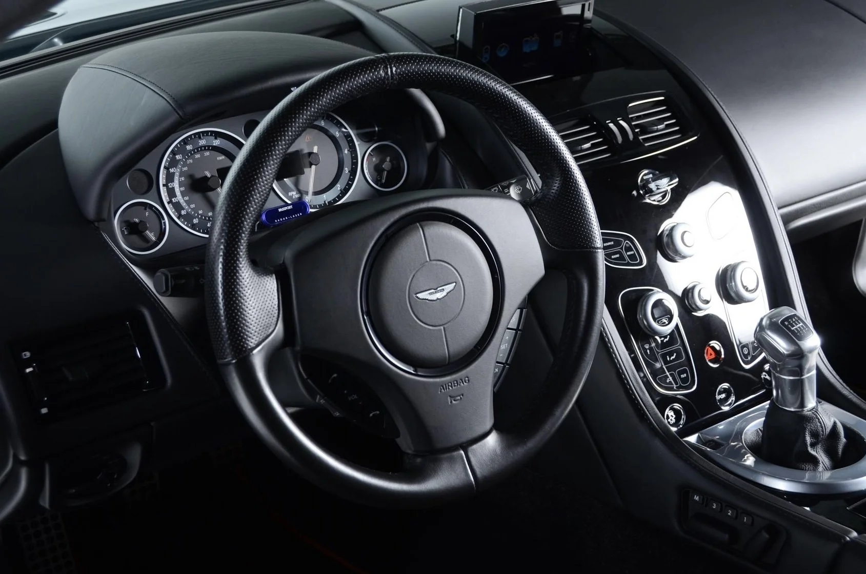 Interior view of a luxury car dashboard and steering wheel, featuring analog gauges, a gear shifter, and control buttons.