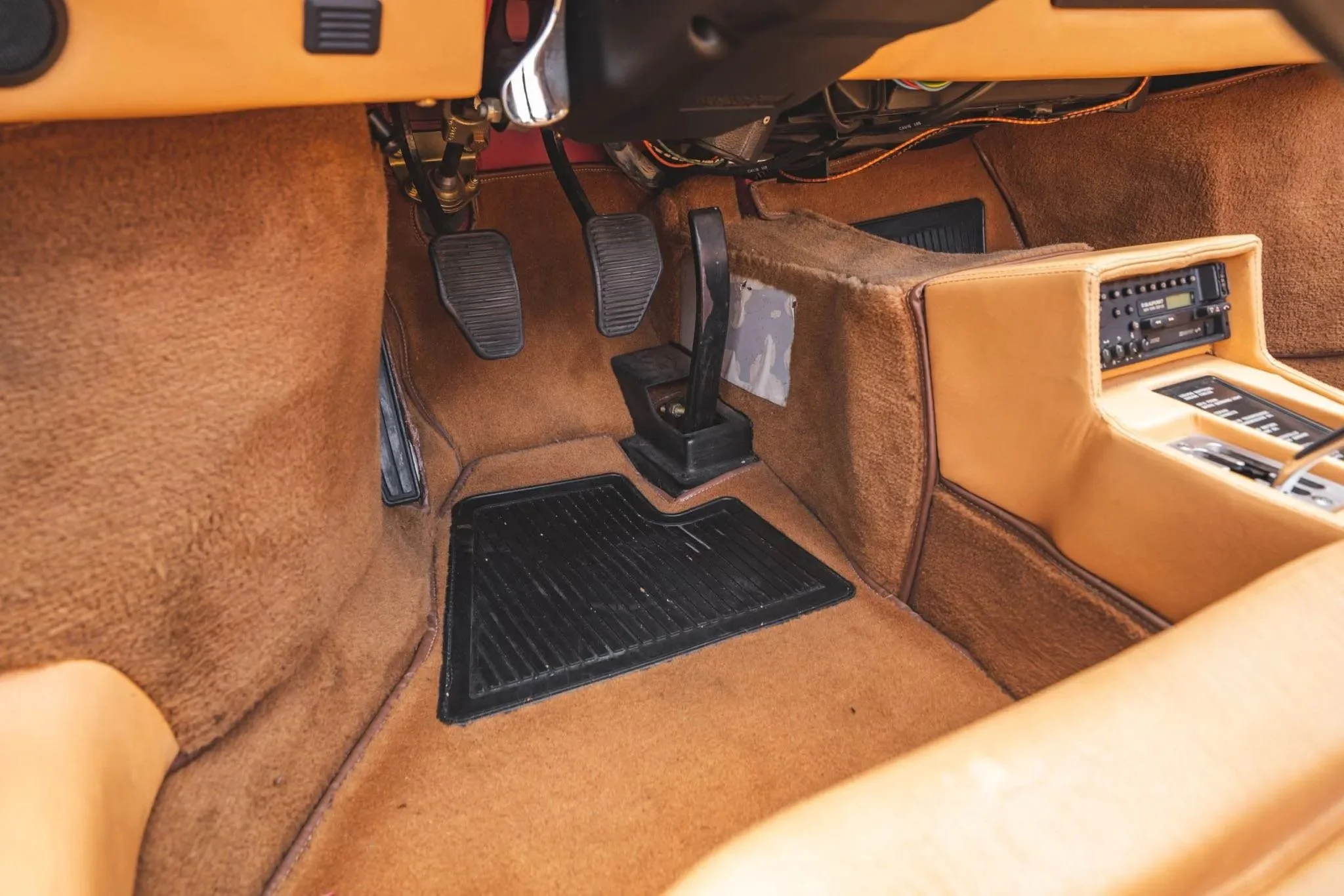 Interior of a vintage car showing the driver's side with accelerator, brake, and clutch pedals, a gear shift in the center, and a dashboard with radio controls, all with tan upholstery and carpeting.