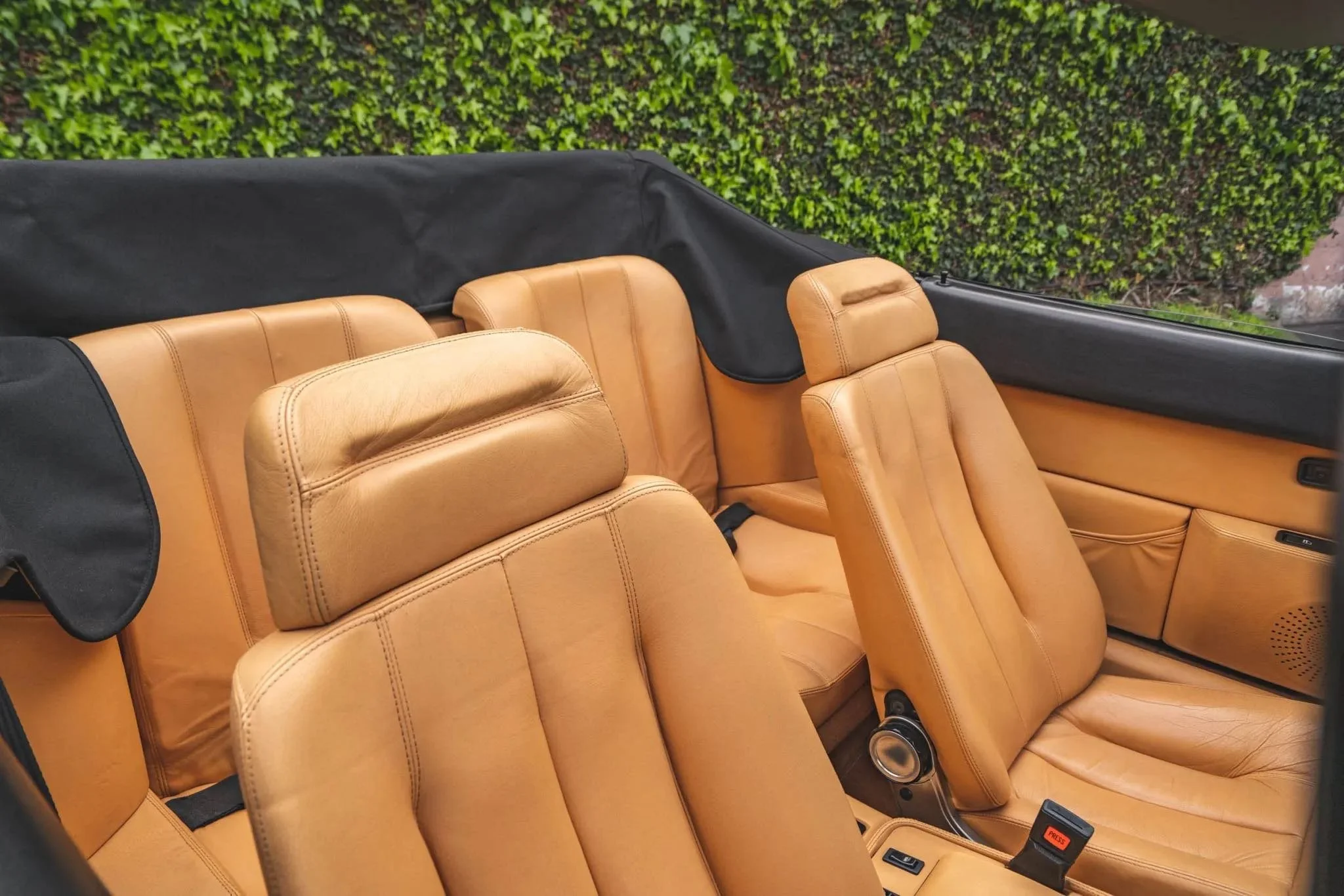 Tan leather car seats inside vintage convertible with a black interior covering and green hedge background.