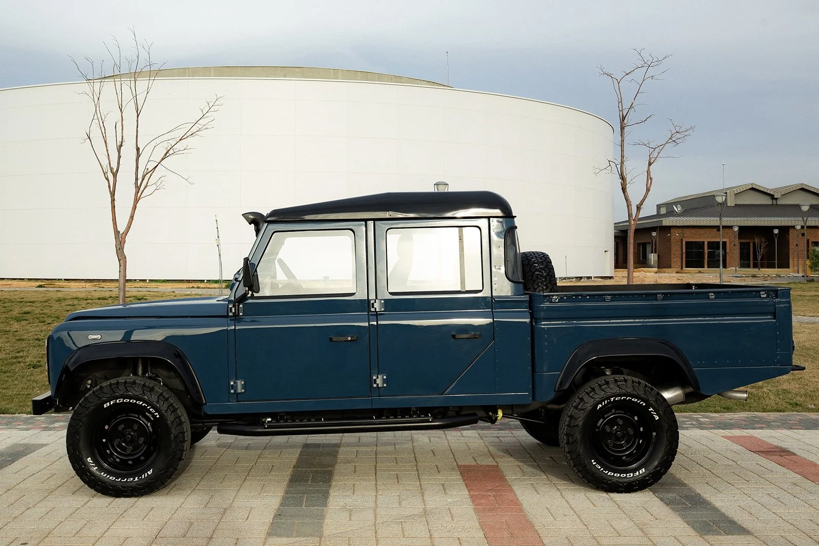 A vintage dark blue pickup truck with a black roof parked on a brick-paved area, with bare trees and a modern white building in the background.