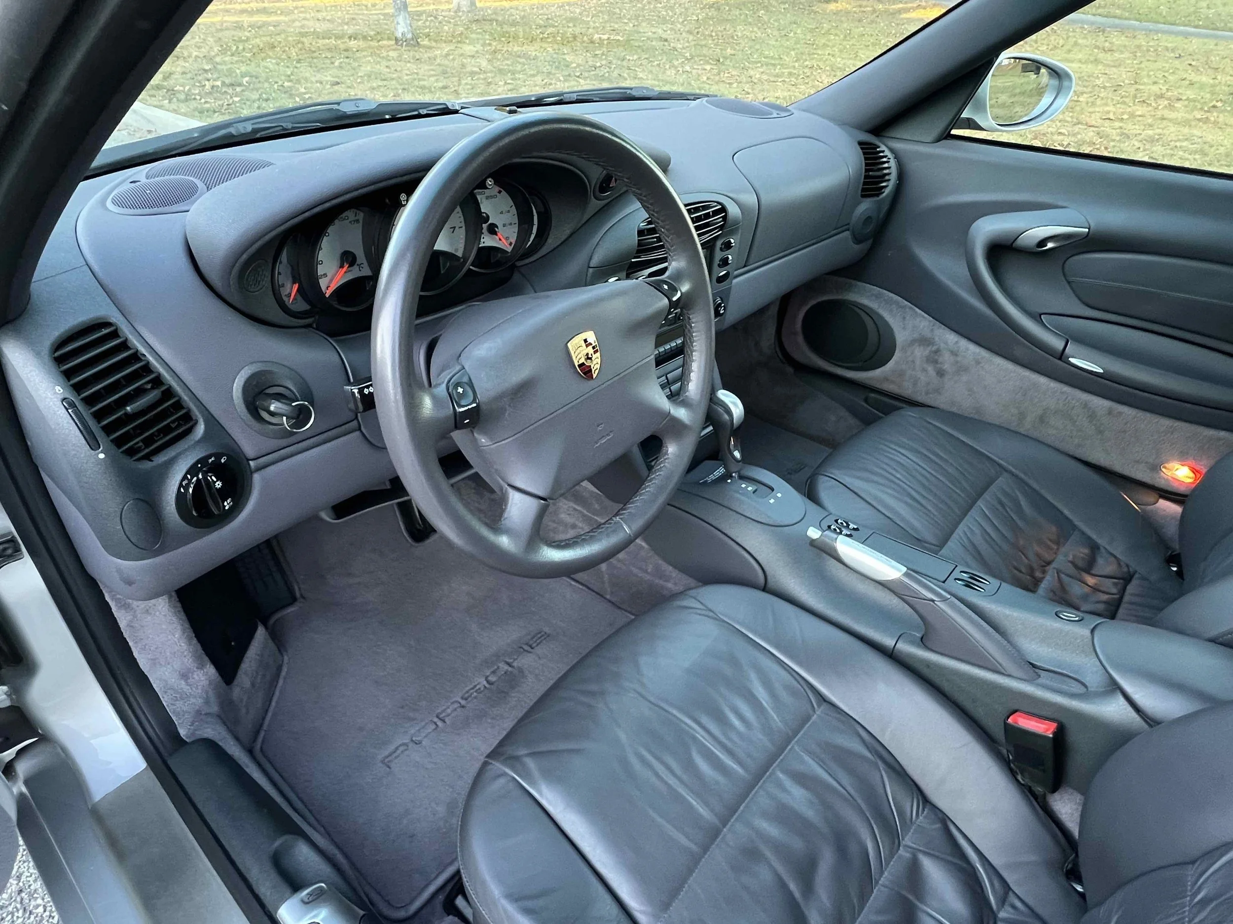 The interior of a Porsche sports car, featuring a leather steering wheel with the Porsche crest, an instrument cluster with a speedometer and tachometer, a central console with controls, black leather seats, and a gray dashboard.