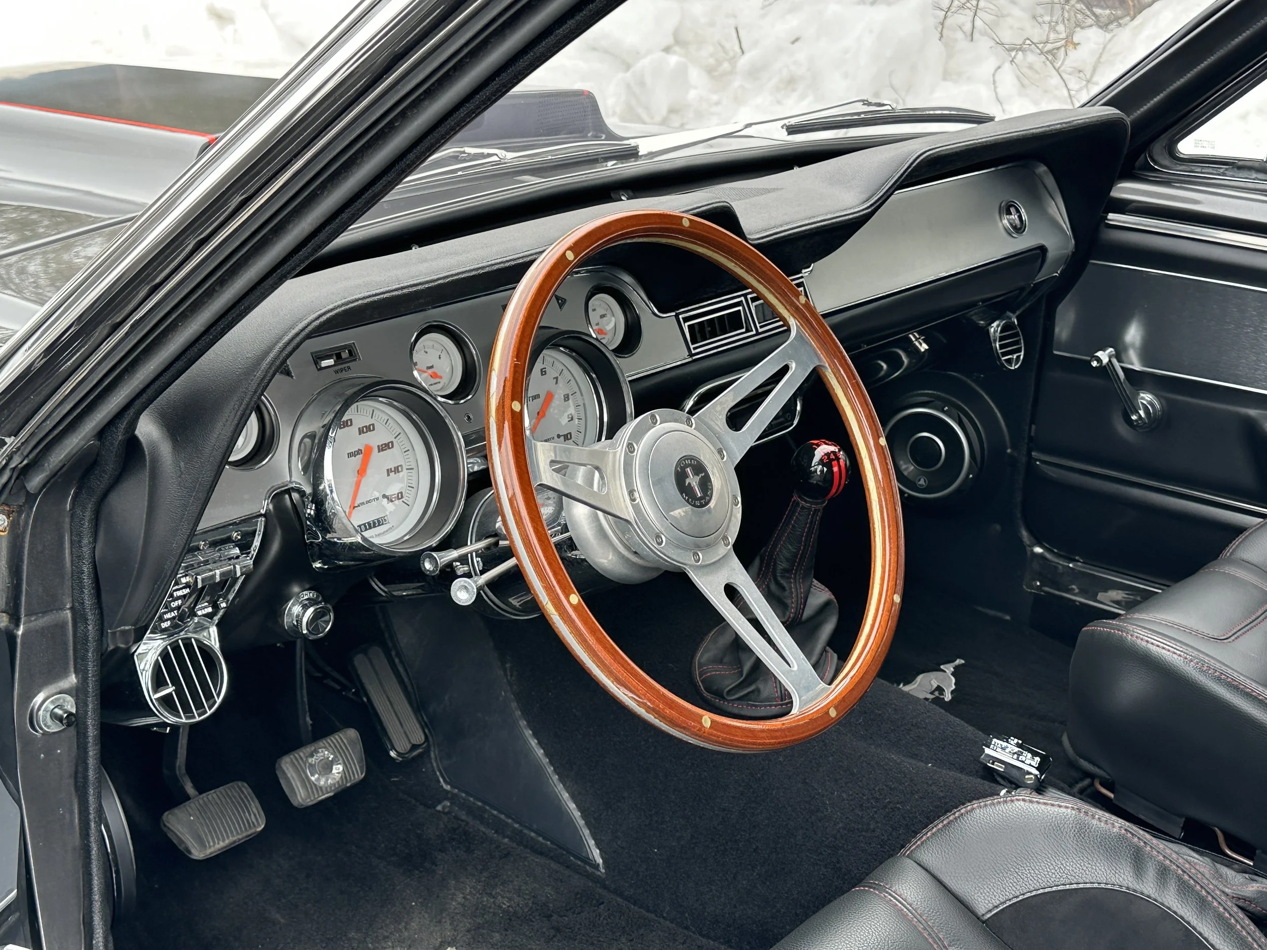 Interior of a vintage Ford Mustang car showing a wooden steering wheel, black dashboard with various gauges, and manual transmission gear shift.