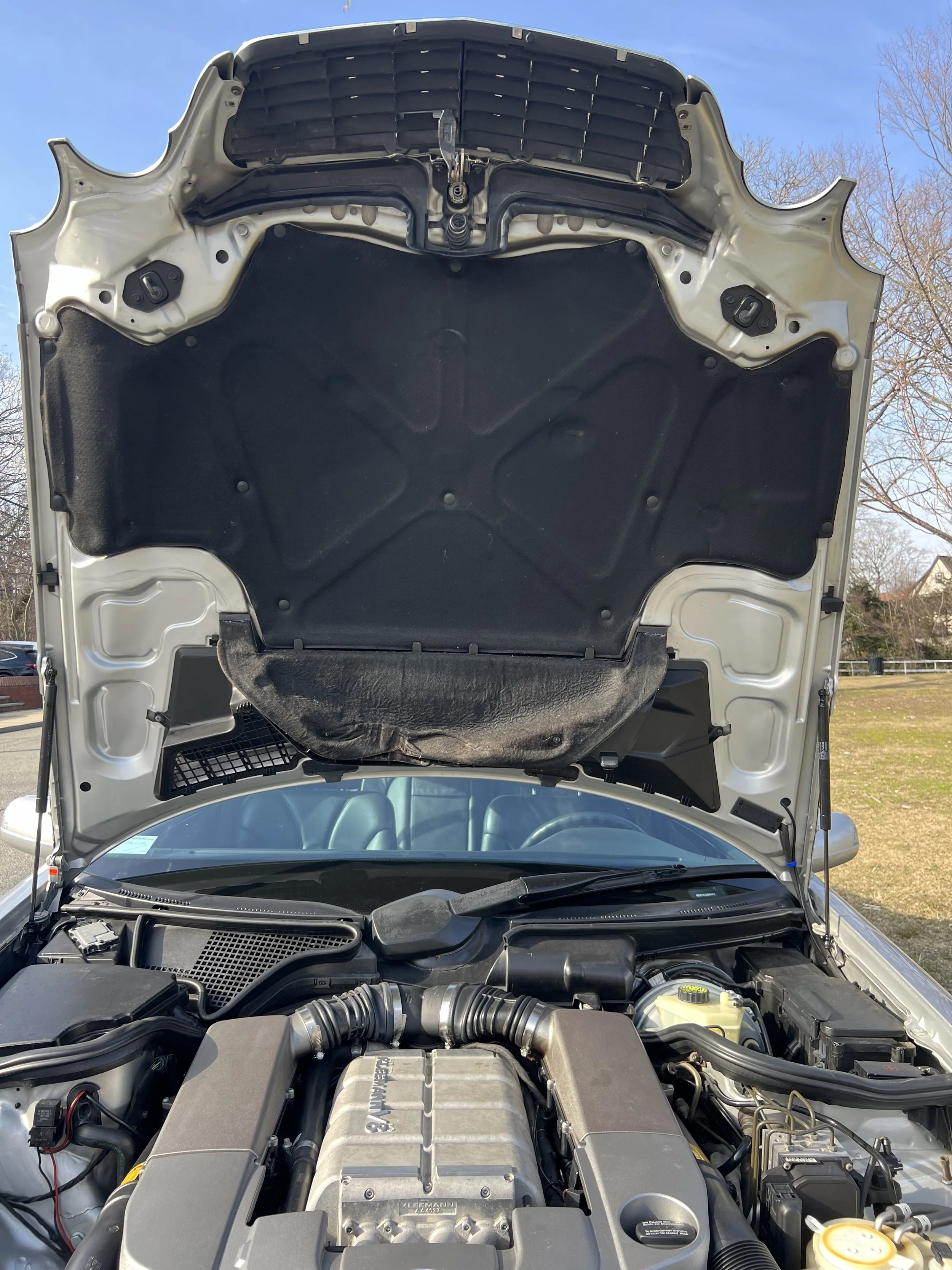 Open hood of a car showing the engine bay with various components, hoses, and the engine cover, with a blue sky and trees in the background.