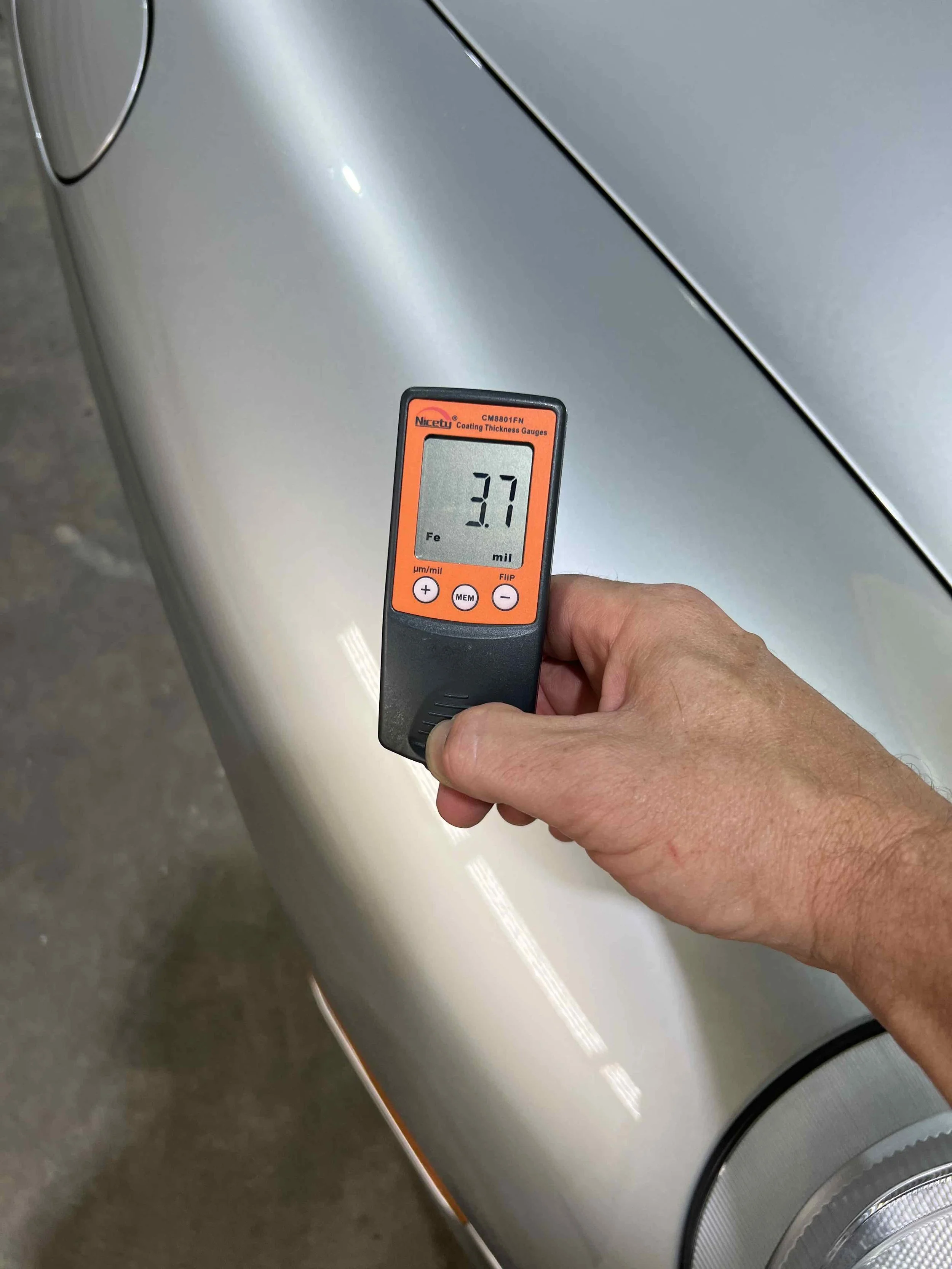 Hand holding a coating thickness gauge close to a silver car's body, displaying a measurement of 3.7 mil.