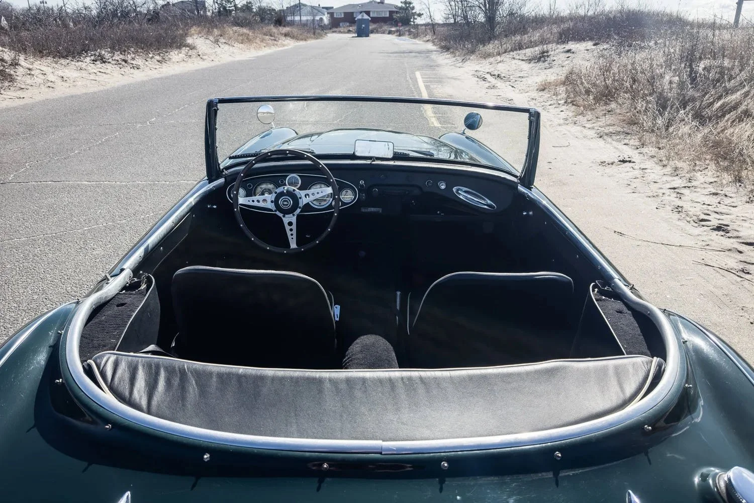 The interior of a vintage sports car, showing the steering wheel, dashboard, and two front seats, with a sandy roadside and a house in the background.