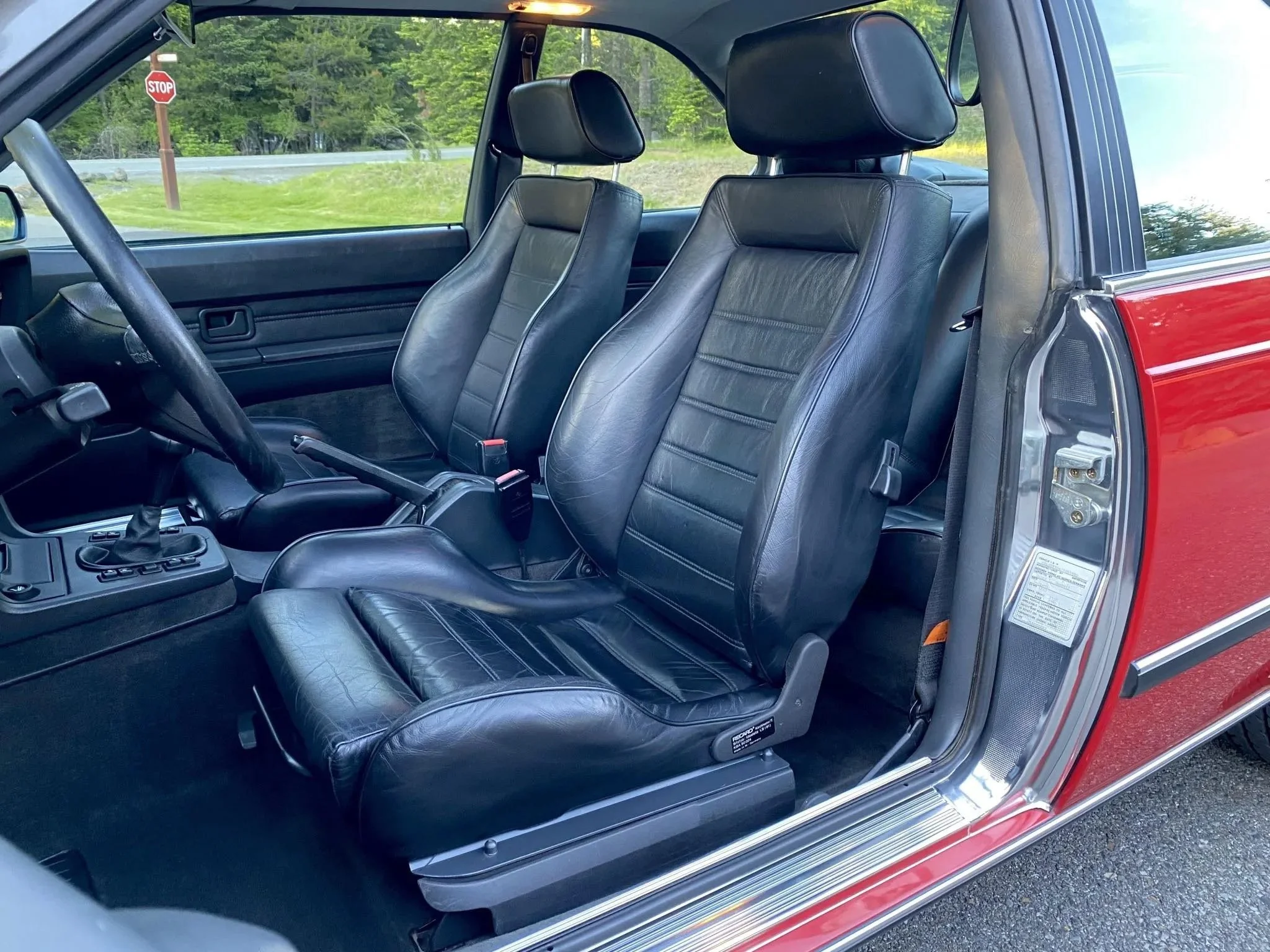 Front interior of a classic red car, showing black leather seats with headrests, a black dashboard, and a steering wheel.
