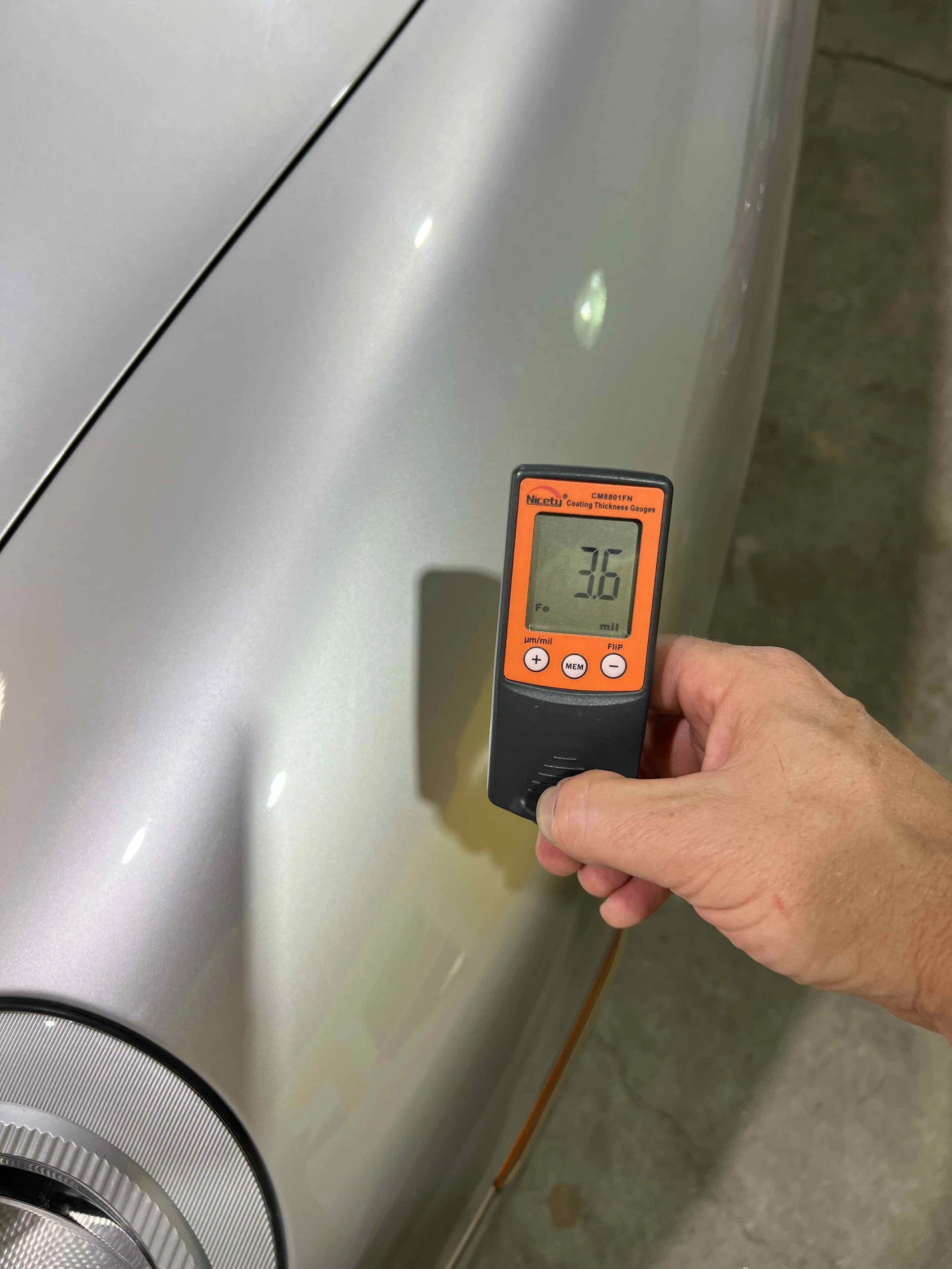 A hand holding a thickness gauge measuring the coating thickness on a silver car's surface, displaying a reading of 3.6 micrometers.