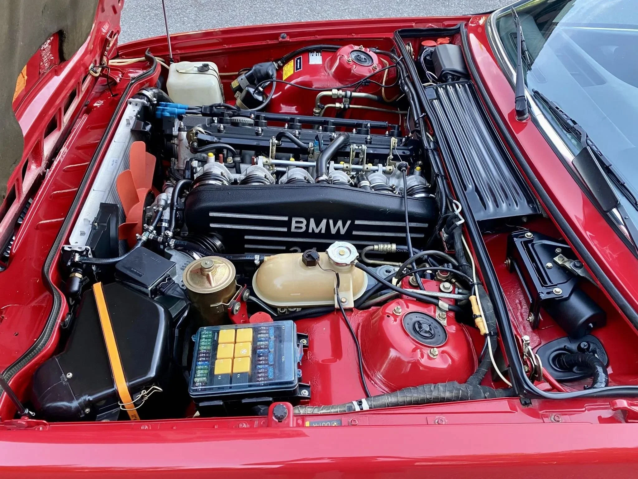 View of a car's engine bay, featuring a red exterior, with an engine labeled BMW, surrounded by various mechanical components such as a coolant reservoir, fuse box, and electrical wiring.