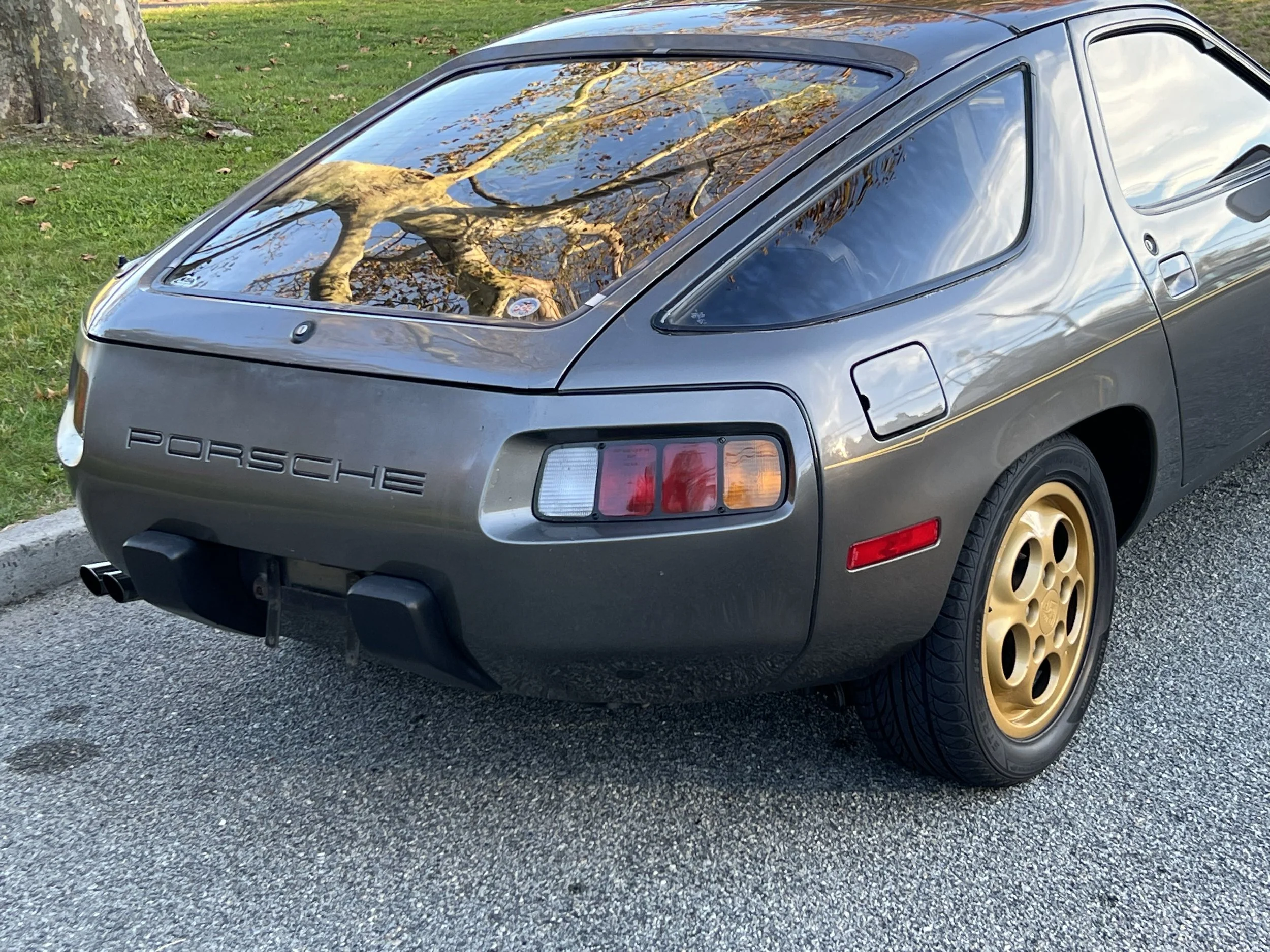 Rear view of a vintage gray Porsche 911 sports car with gold wheels, parked on a paved street beside a grassy area with trees.