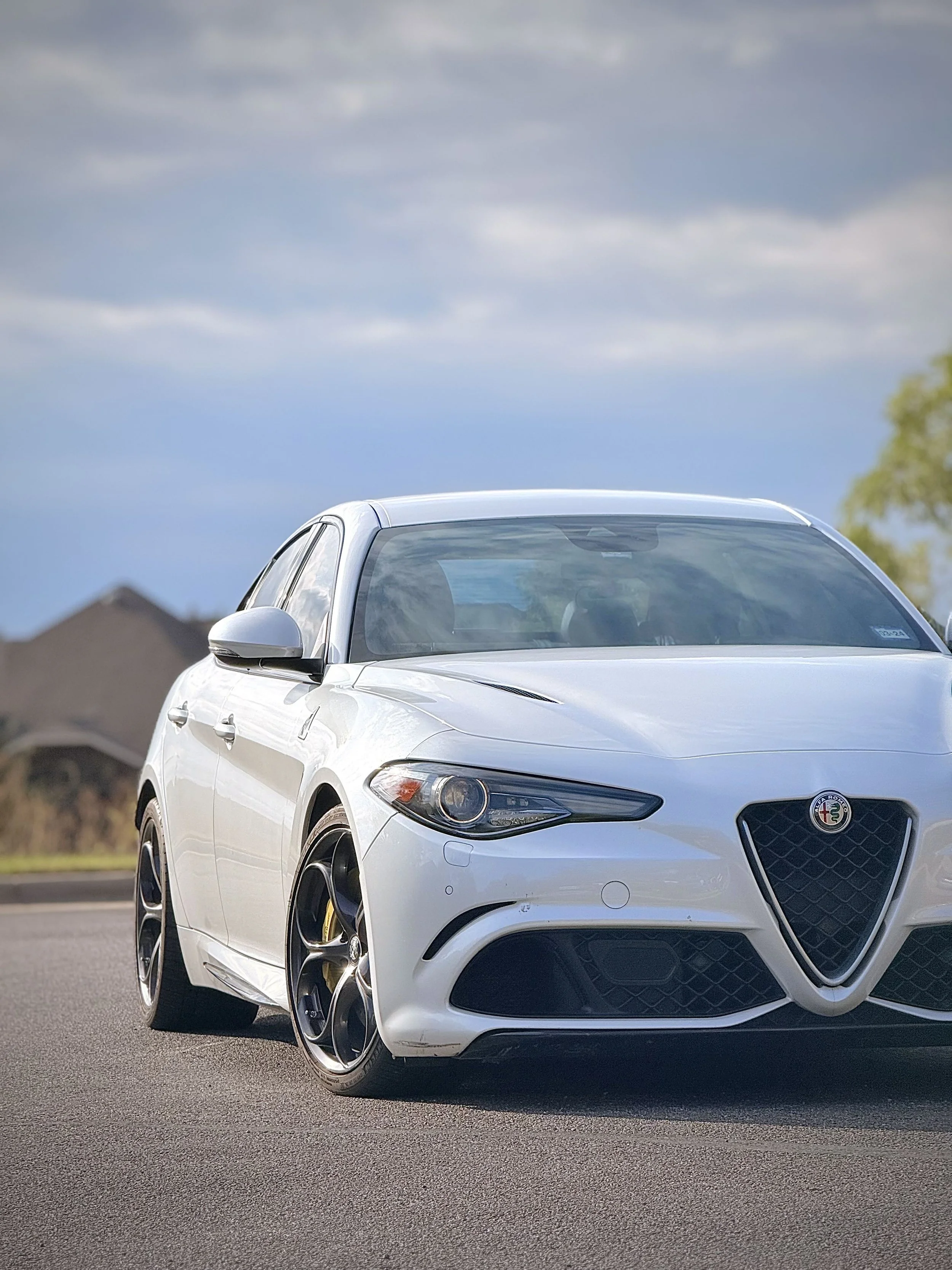 A white Alfa Romeo sports car on a paved road with a cloudy sky and trees in the background.