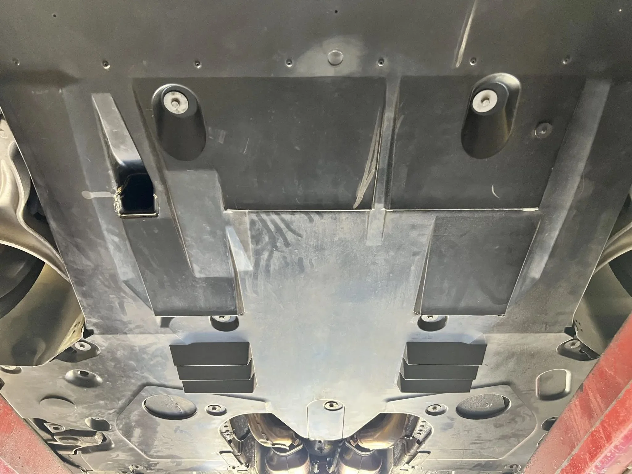Underbody of a vehicle showing metal surface, bolts, and plastic components, likely part of the chassis or undercarriage.