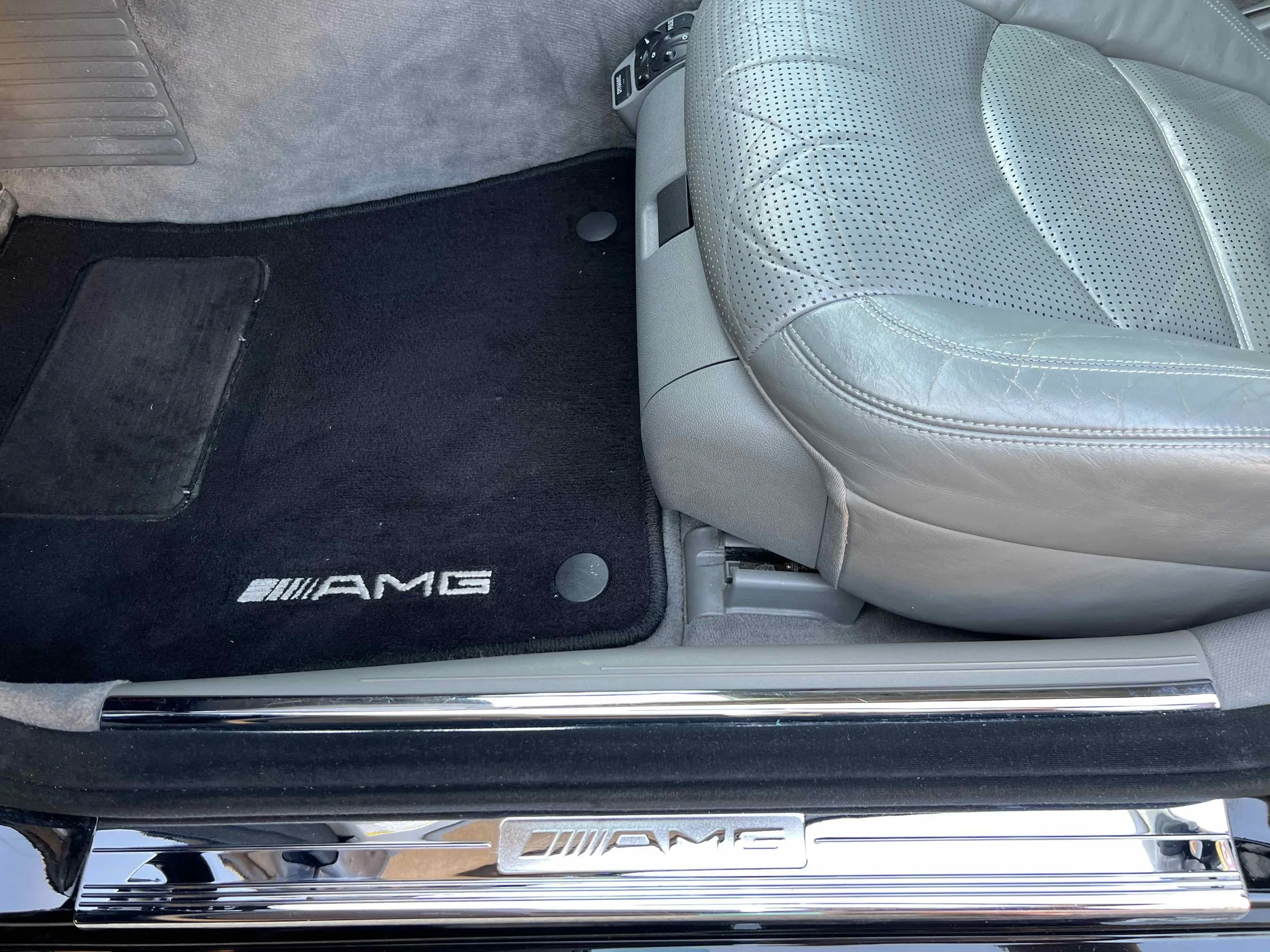 Front passenger side of a Mercedes-Benz AMG vehicle interior, showing a gray leather seat, black AMG floor mat, and AMG branded door sill.