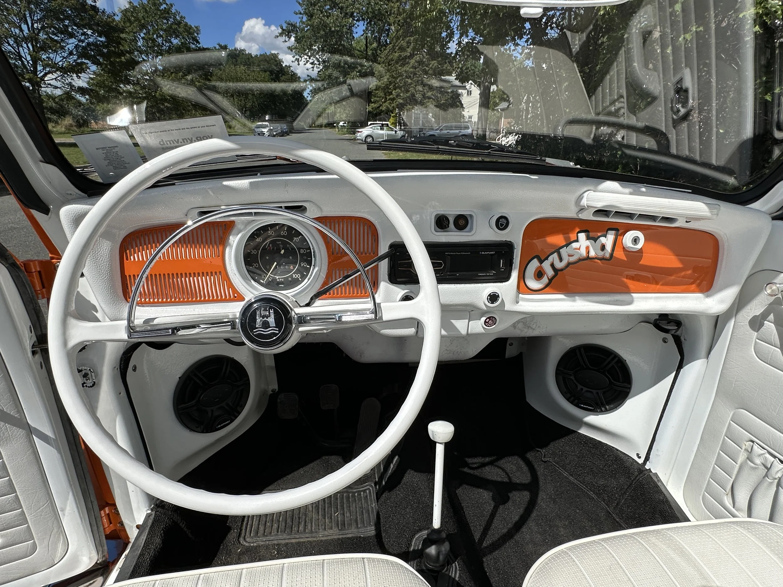 The image shows the dashboard of a vintage white and orange Volkswagen vehicle with the word "crushd" on the passenger side. It includes an old-fashioned speedometer, a steering wheel, two speakers, and a gear shift in the center.