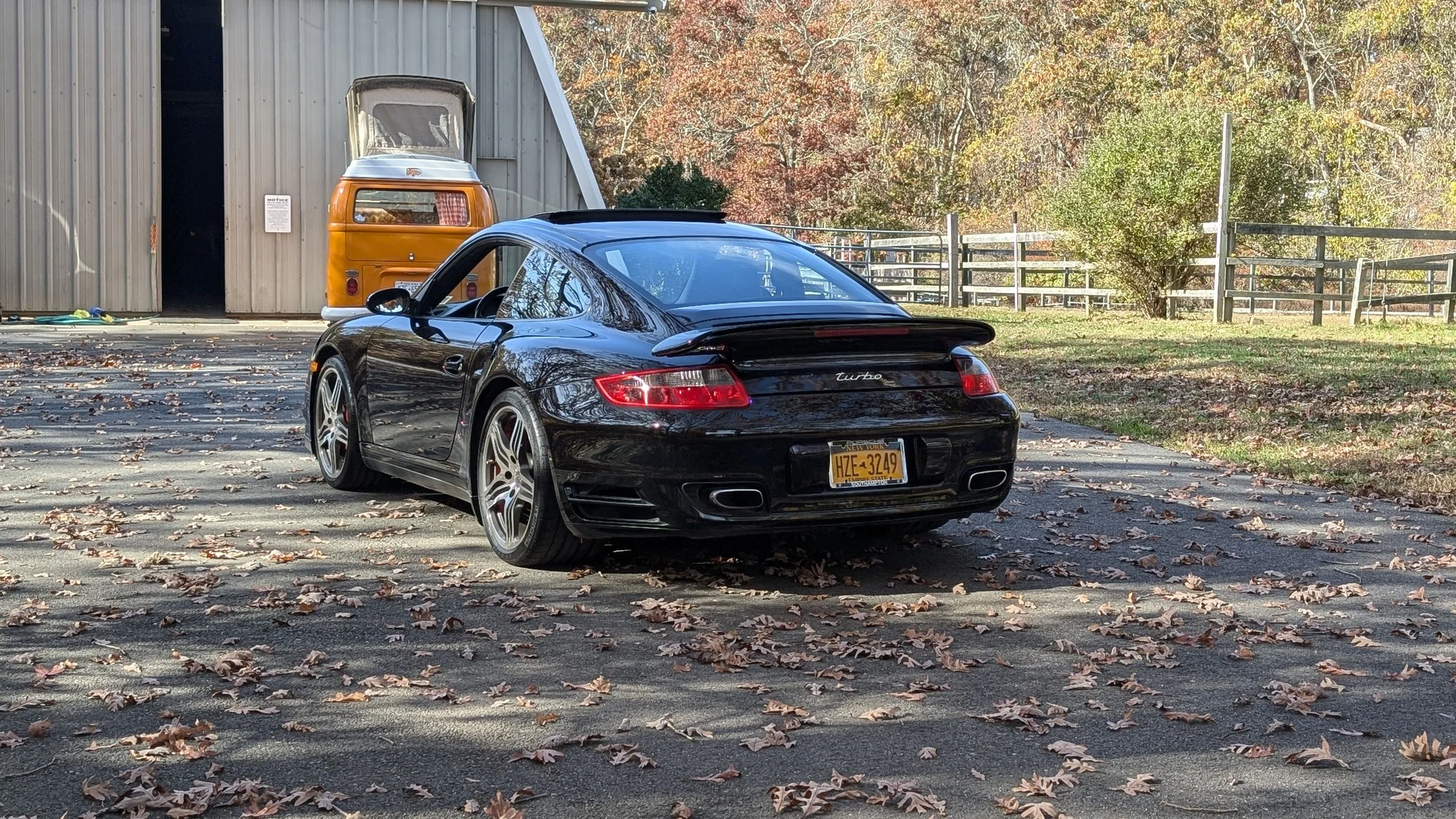 A black Porsche Carrera with a rear spoiler parked on a driveway covered with fallen autumn leaves, near a gray metal barn and an orange vintage van.