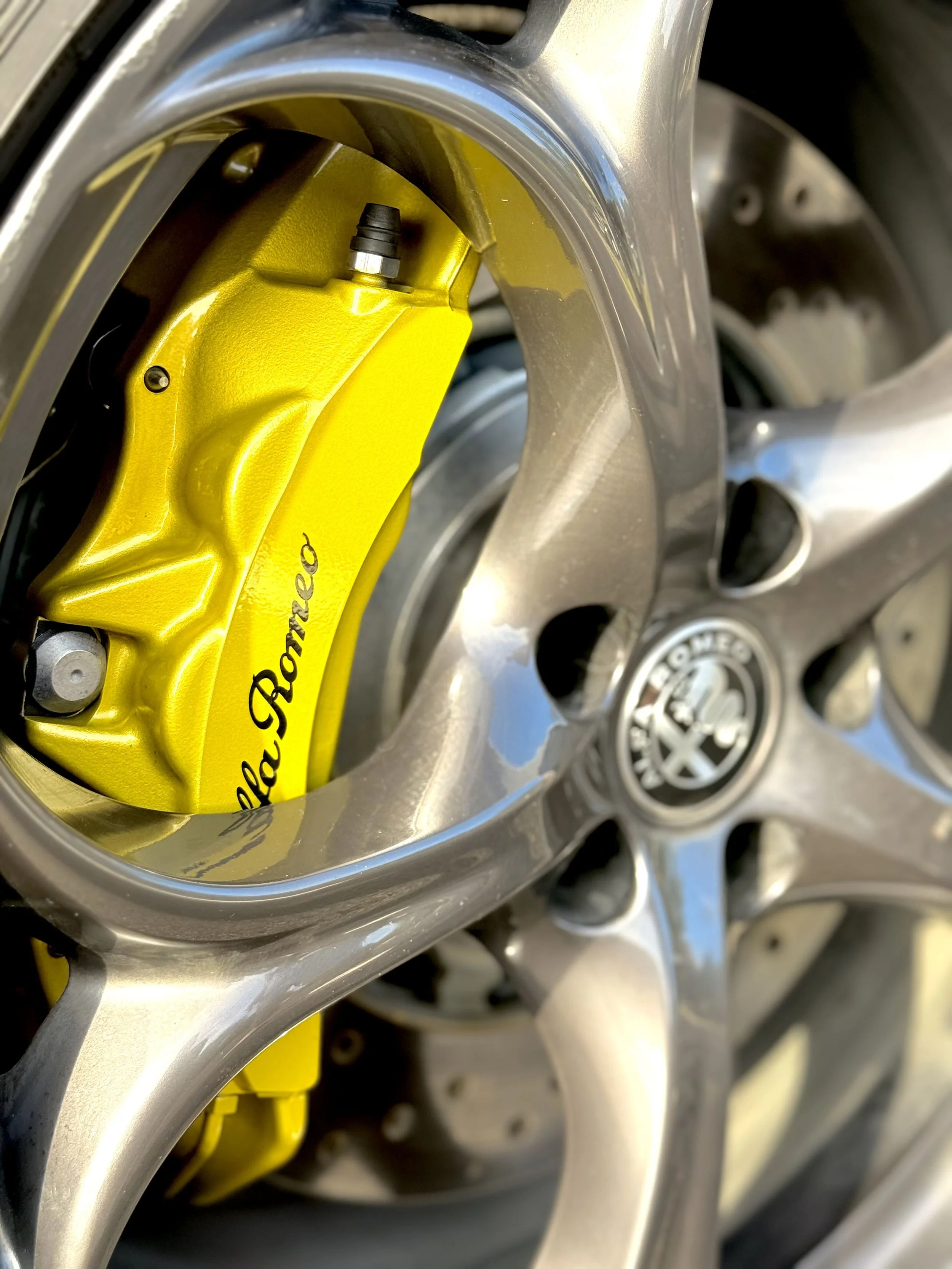 Close-up of a yellow brake caliper with the words 'Alfa Romeo' printed on it, installed on a silver alloy wheel of an Alfa Romeo car.