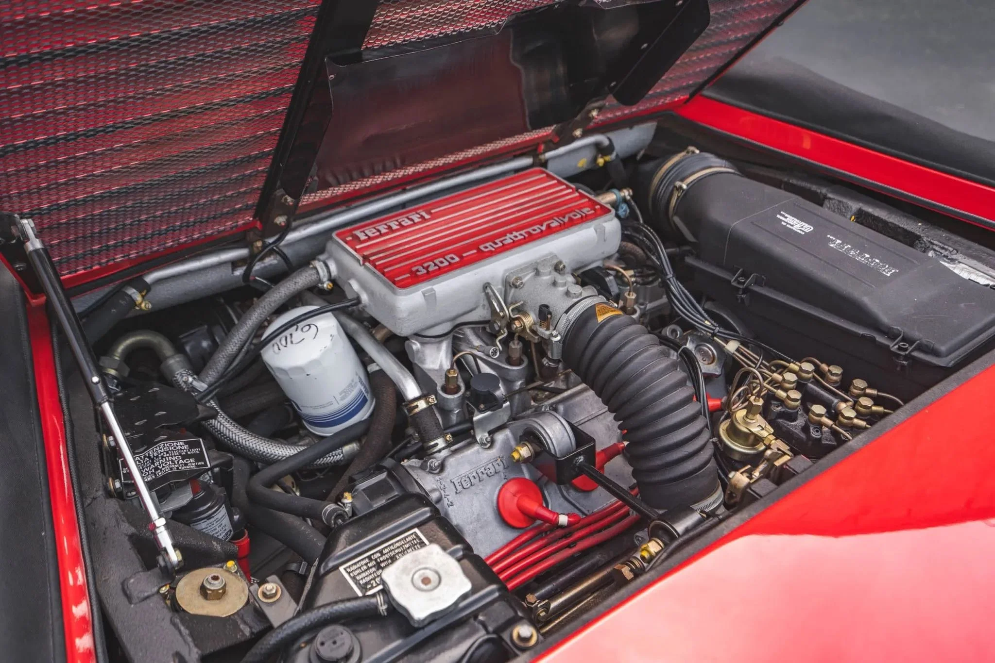Engine bay of a Ferrari sports car with a red intake manifold labeled 'Ferrari 3200 = Quadralvalve' and various mechanical components.
