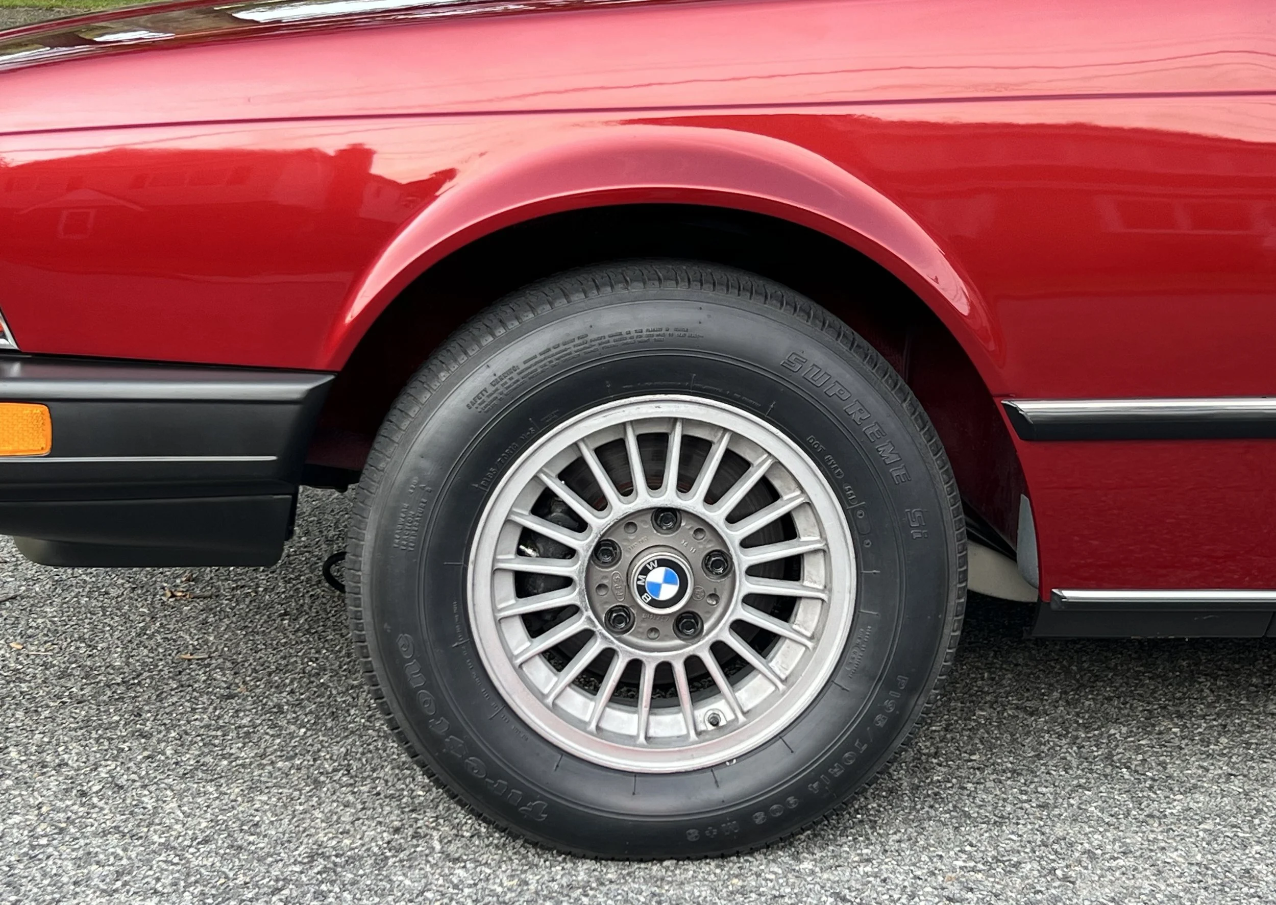 Close-up of a vintage red BMW BMW 630 csi wheel with a alloy rim and Goodyear Super Eagle tire, on a gravel surface.