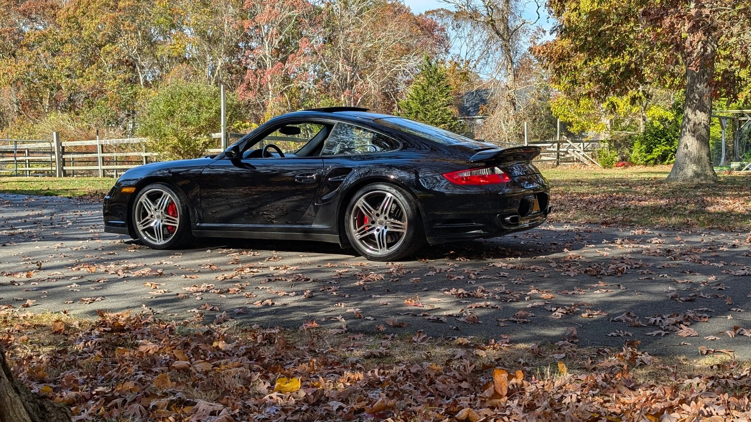 A black Porsche car parked on a driveway covered with fallen autumn leaves, with trees and a wooden fence in the background.