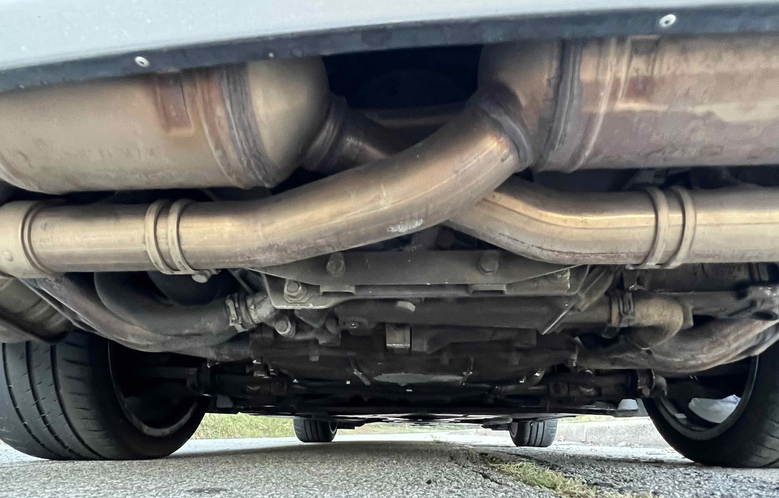 View of the underside of a car showing exhaust pipes, muffler, suspension, and tires on a paved road.