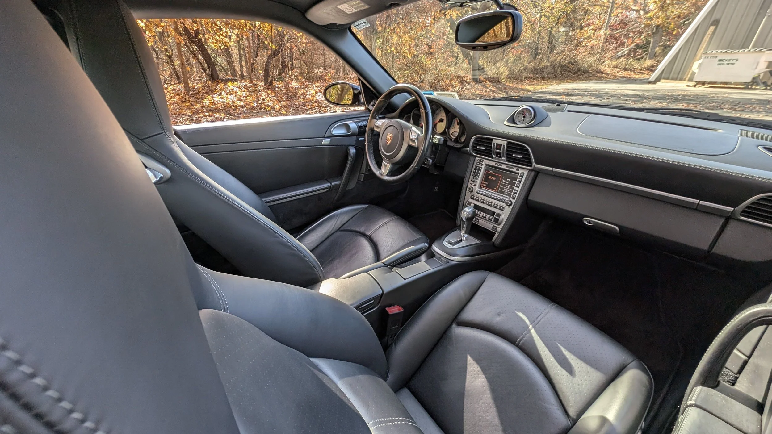 Interior of a black sports car showing the driver and passenger seats, dashboard, steering wheel with Porsche logo, center console with gear shift, and an analog clock on the dashboard. The car is parked outdoors with autumn leaves and trees visible 