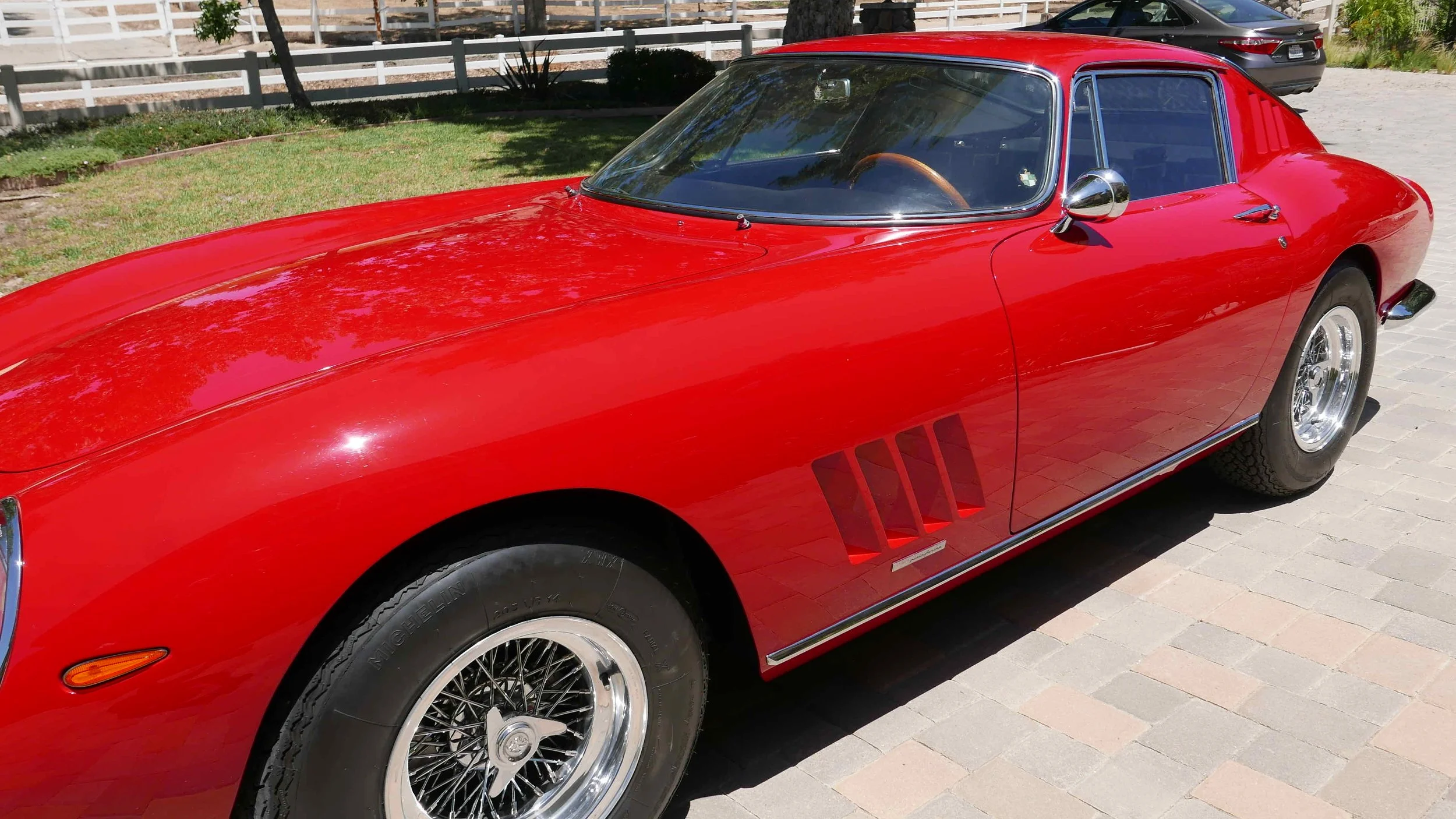 Red vintage sports car parked on a paved area with green grass and trees in the background.