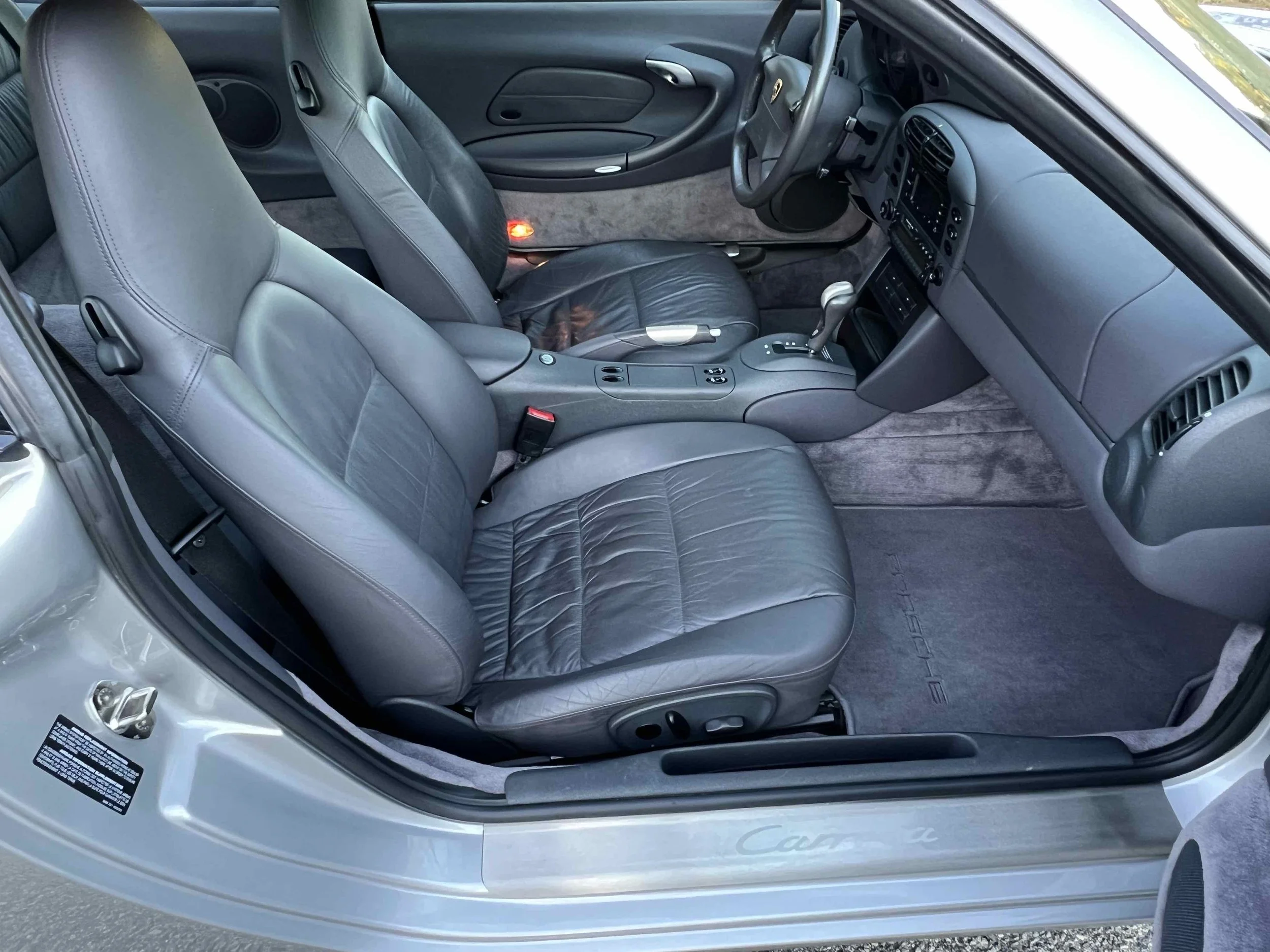 Interior of a gray Porsche sports car, showing two leather front seats, gray dashboard, steering wheel, gear shift, center console, and gray carpeted floor mats.