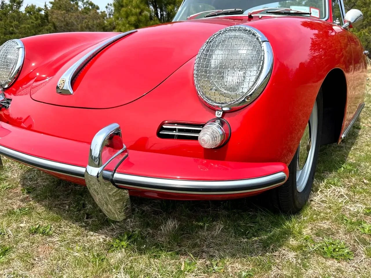 Close-up of a classic red Porsche 356 with chrome accents, grille, and protective wire covers on the headlights, parked outdoors on grass.