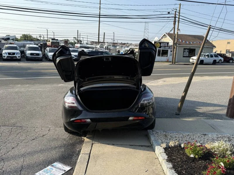 Black car with open trunk parked on sidewalk with a small flower bed nearby, and a busy street with parked and moving cars and utility poles in the background.