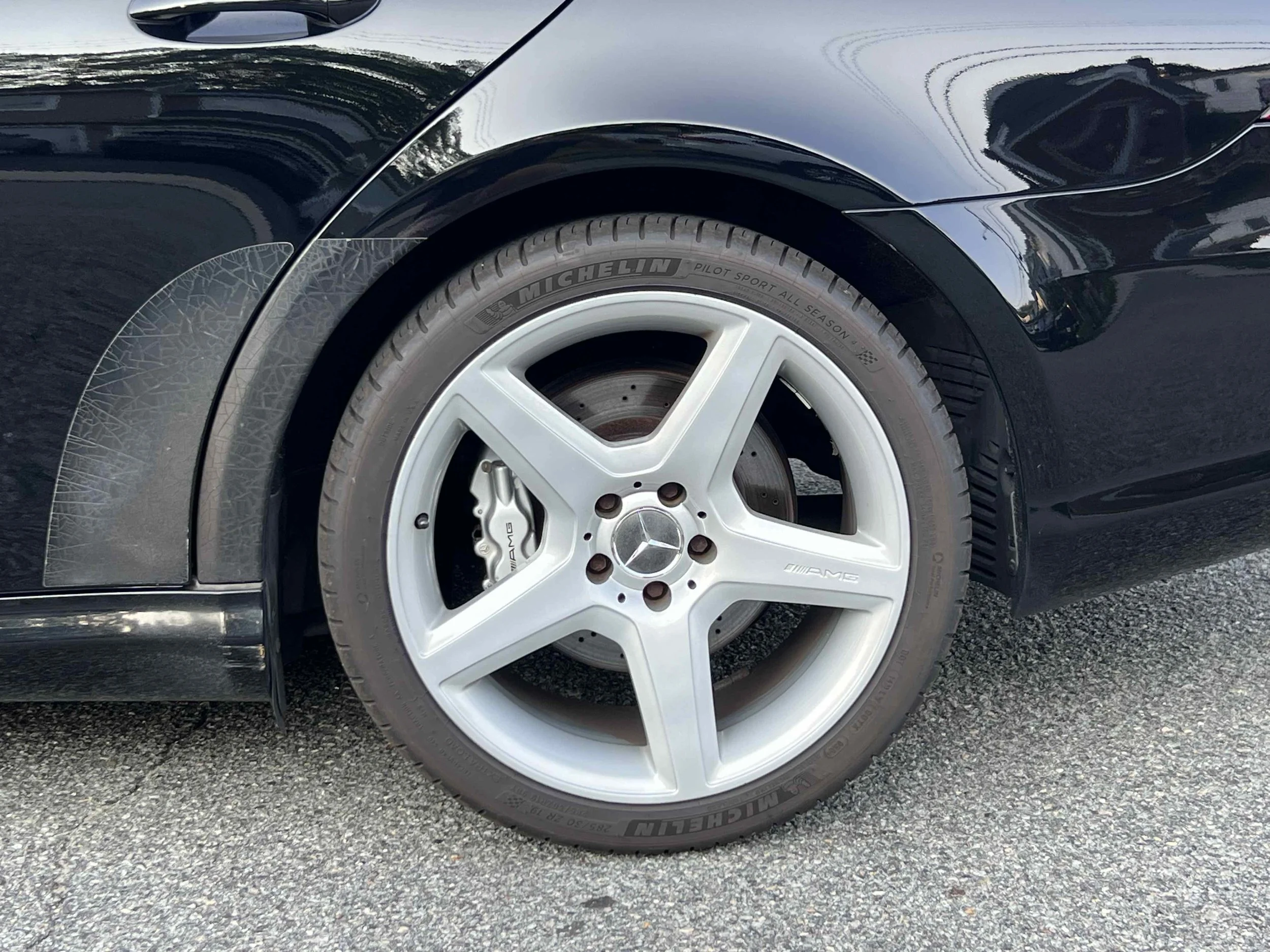 Front tire and wheel of a black Mercedes-Benz car with AMG brake calipers, parked on an asphalt surface.