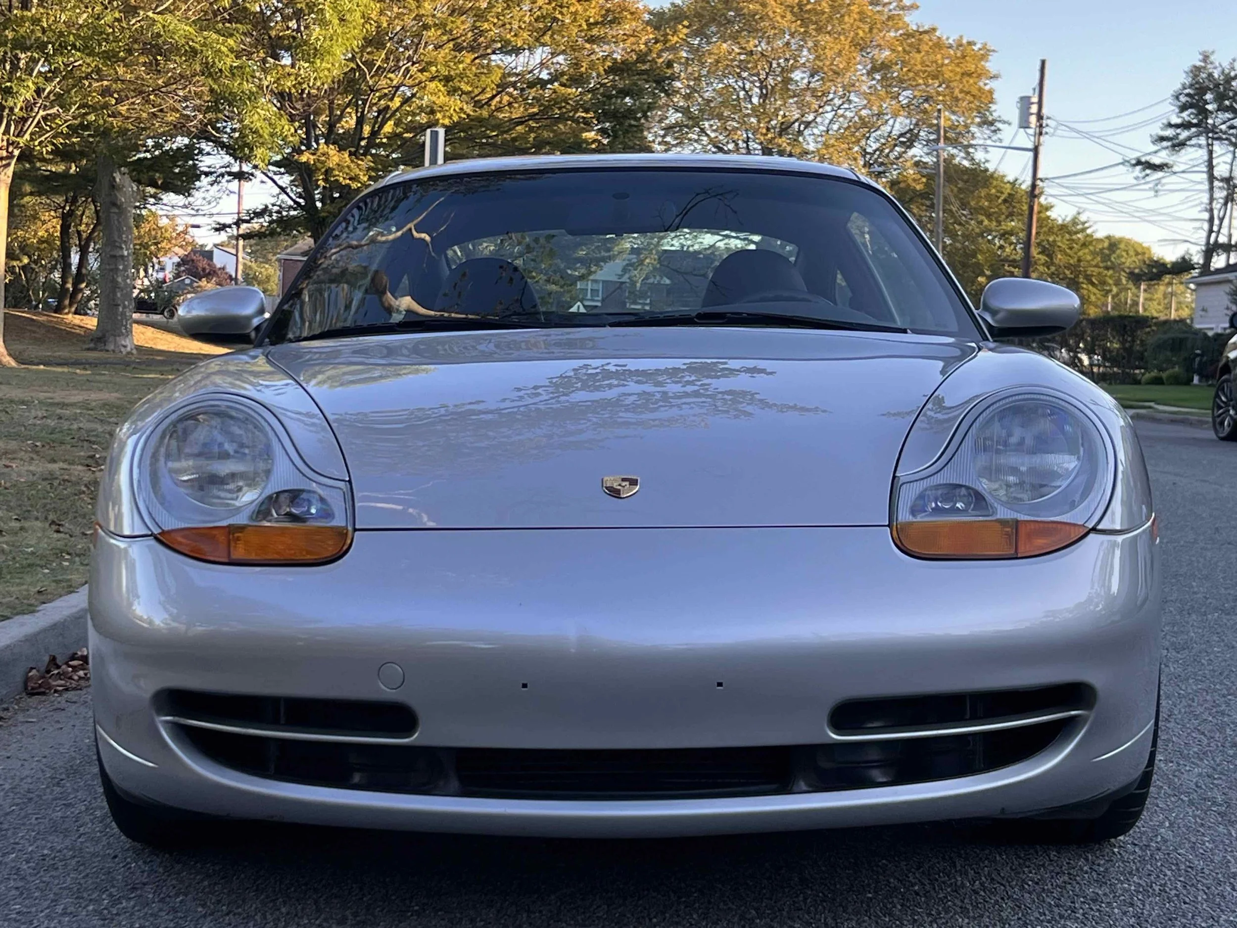 Front view of a silver Porsche sports car parked on a street with trees and houses in the background.