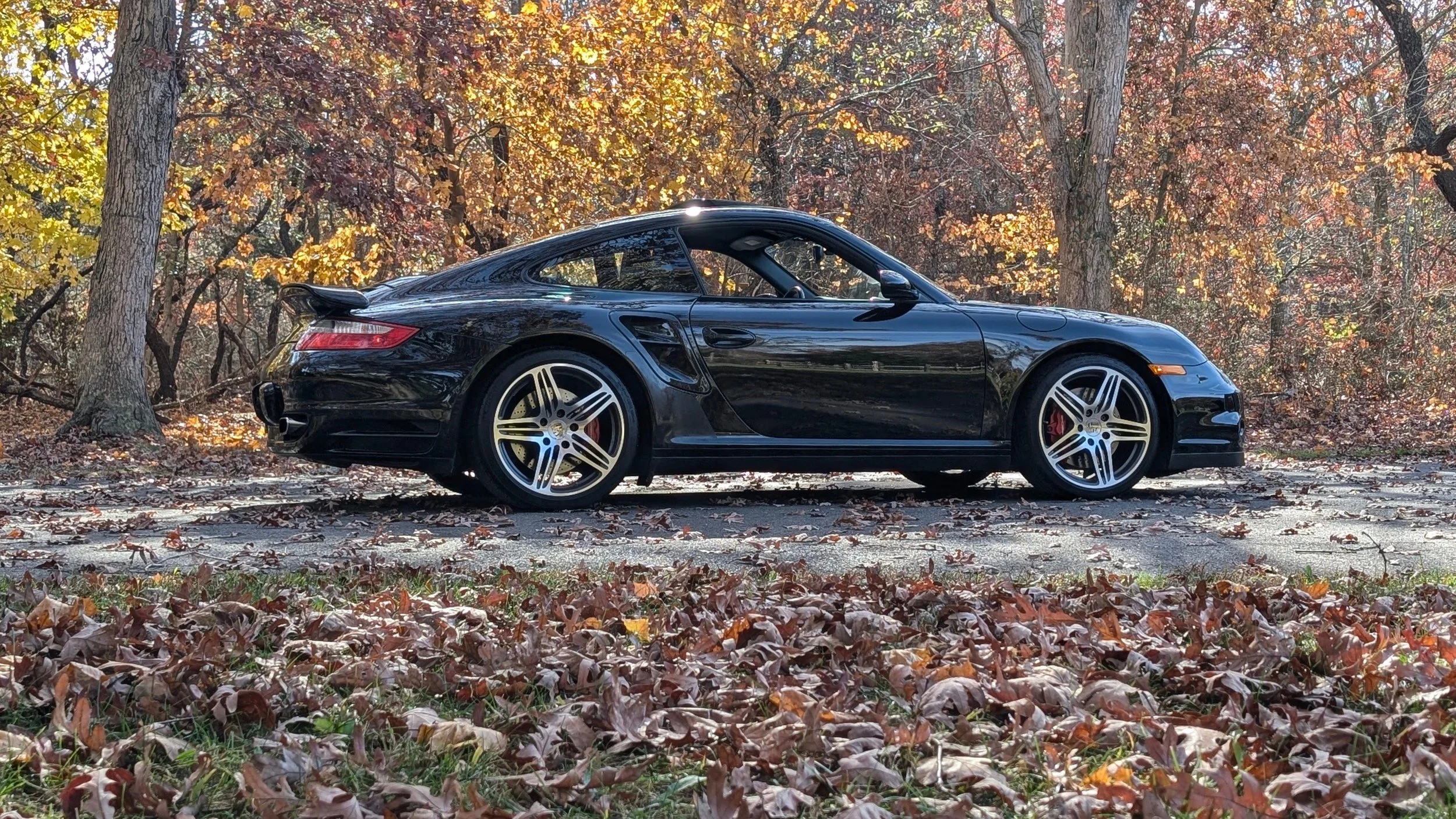 Black sports car parked on a leaf-covered street with autumn trees in the background.
