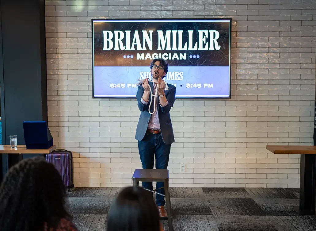 Brian Miller founder and principal consultant of Clarity Up performing on stage in front of a large screen with his name, Brian Miller. The magician is holding a white rope and making a gesture as if explaining a trick.