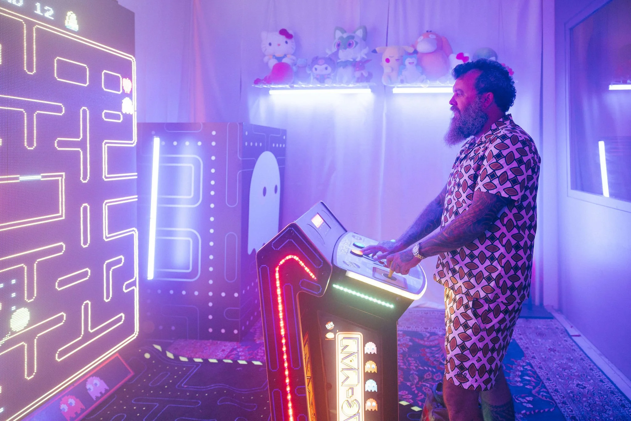A man with tattoos and a beard playing Pac-Man on an arcade machine, with neon lights and Pac-Man-themed decor in the background.