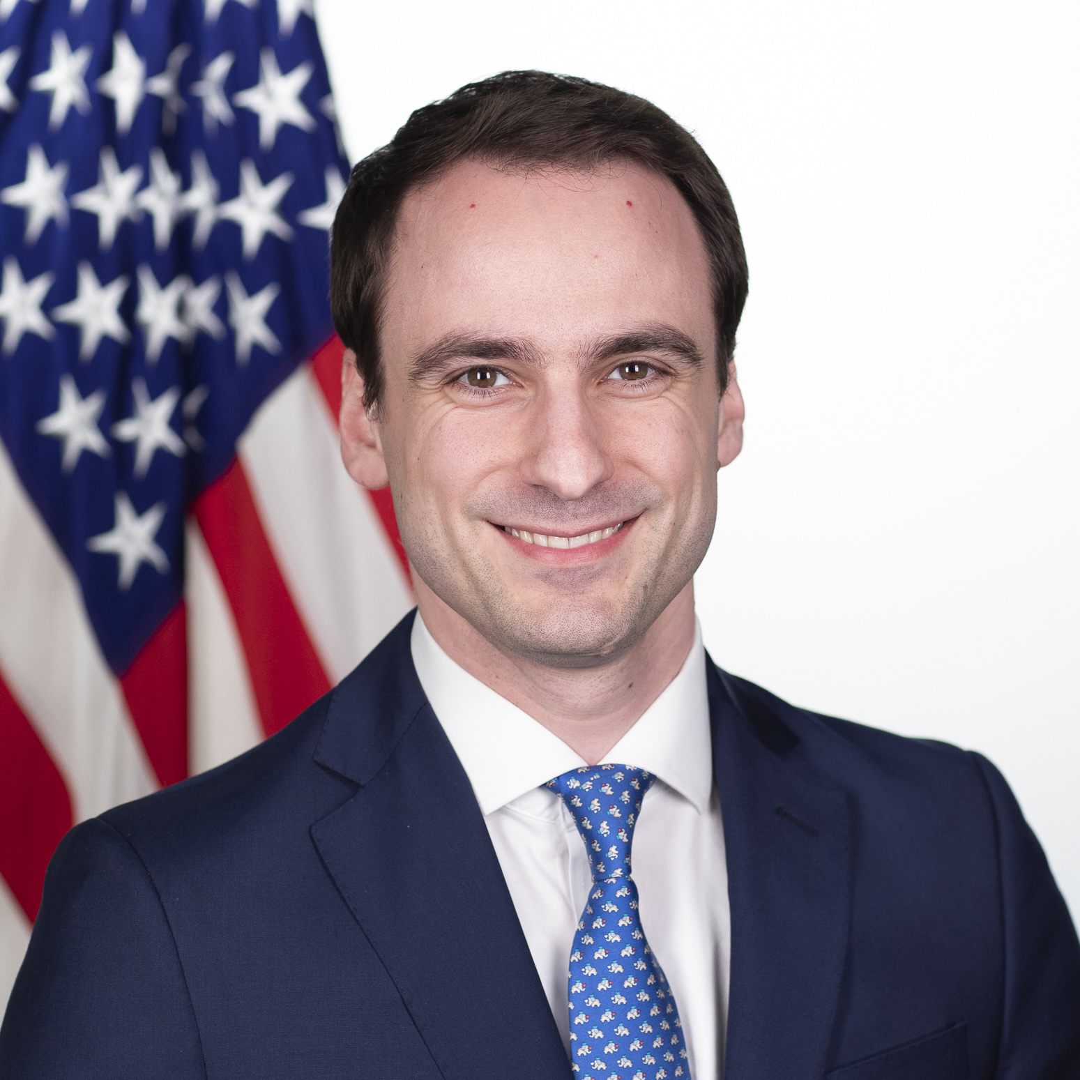 Young man in a navy suit and tie smiling, with an American flag in the background.