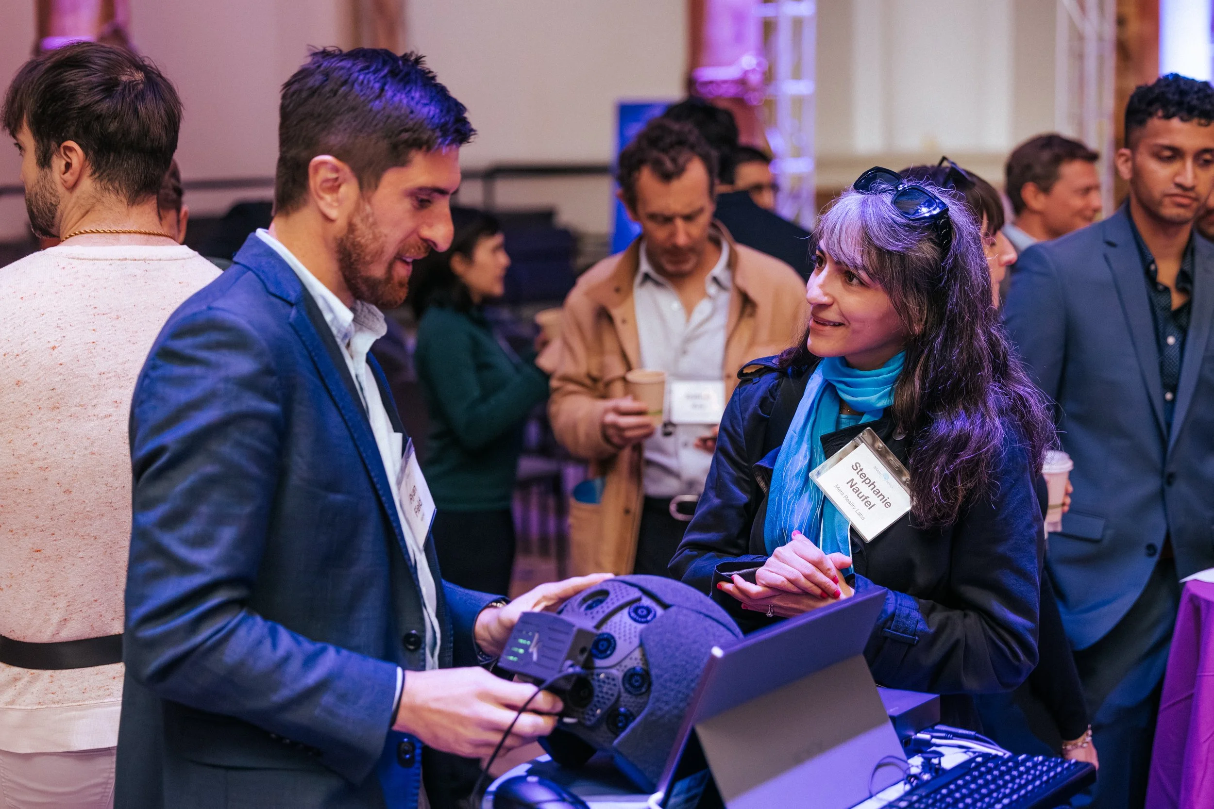 A man showing a device to a woman at a networking event, with other people in the background holding coffee cups.