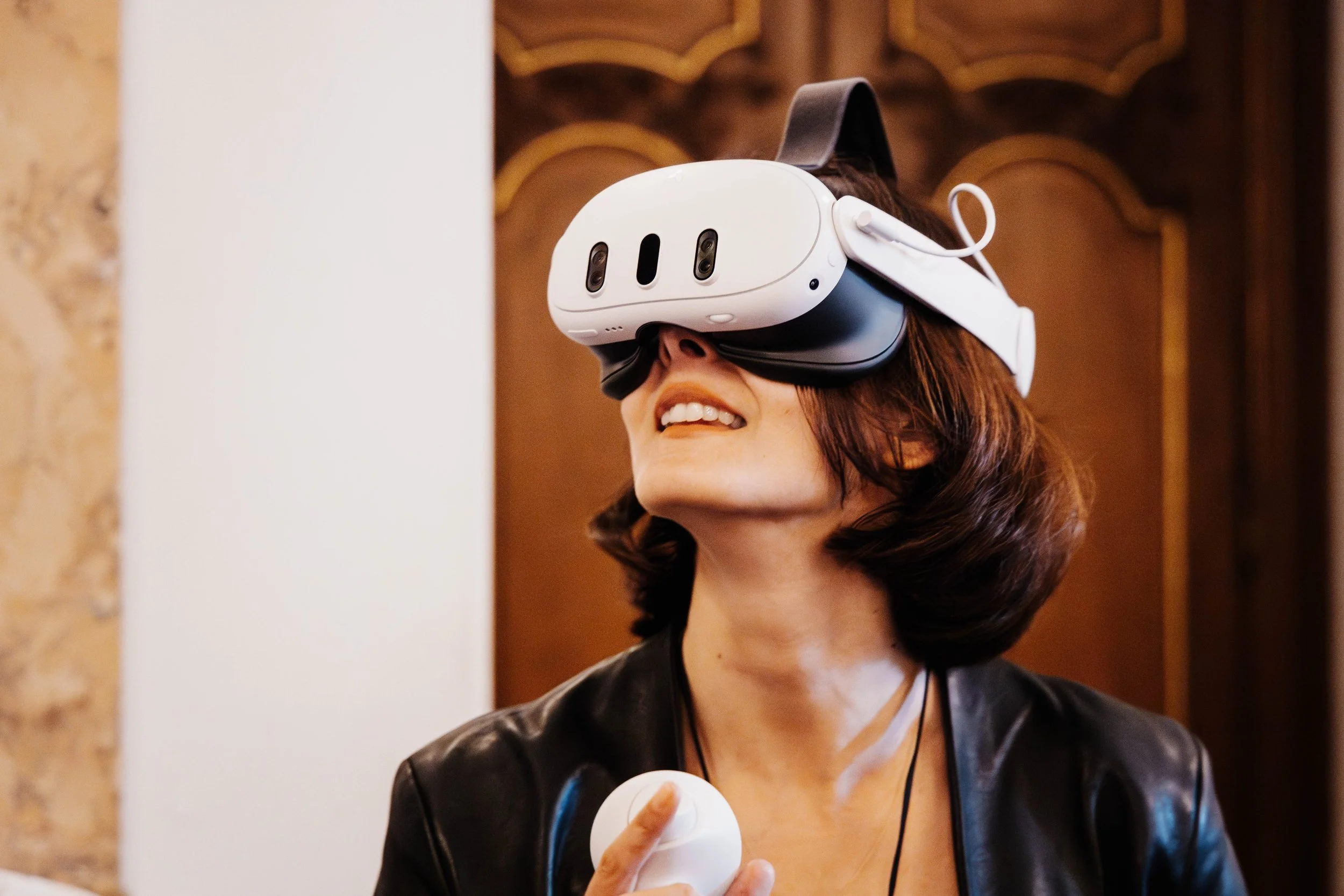 A woman with shoulder-length brown hair using a virtual reality headset and holding a VR controller, smiling in a room with warm lighting.