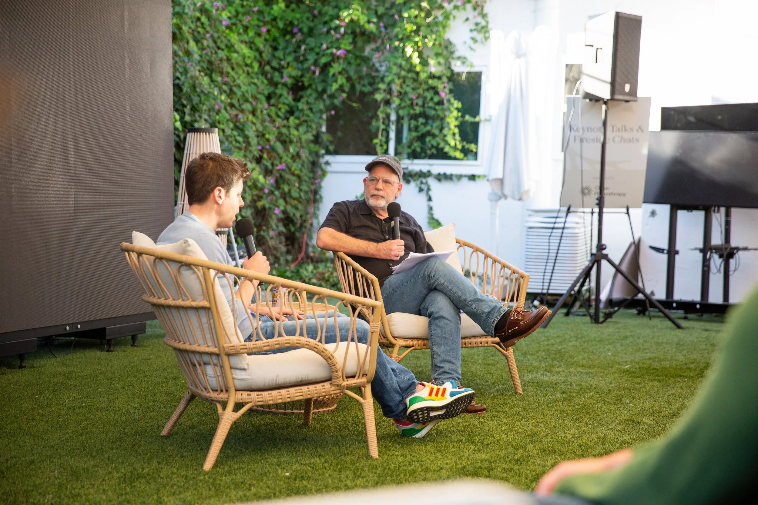 Two men sitting in outdoor wicker chairs having an interview with microphones, one with a laptop, in a garden setting with greenery and a white building in the background.