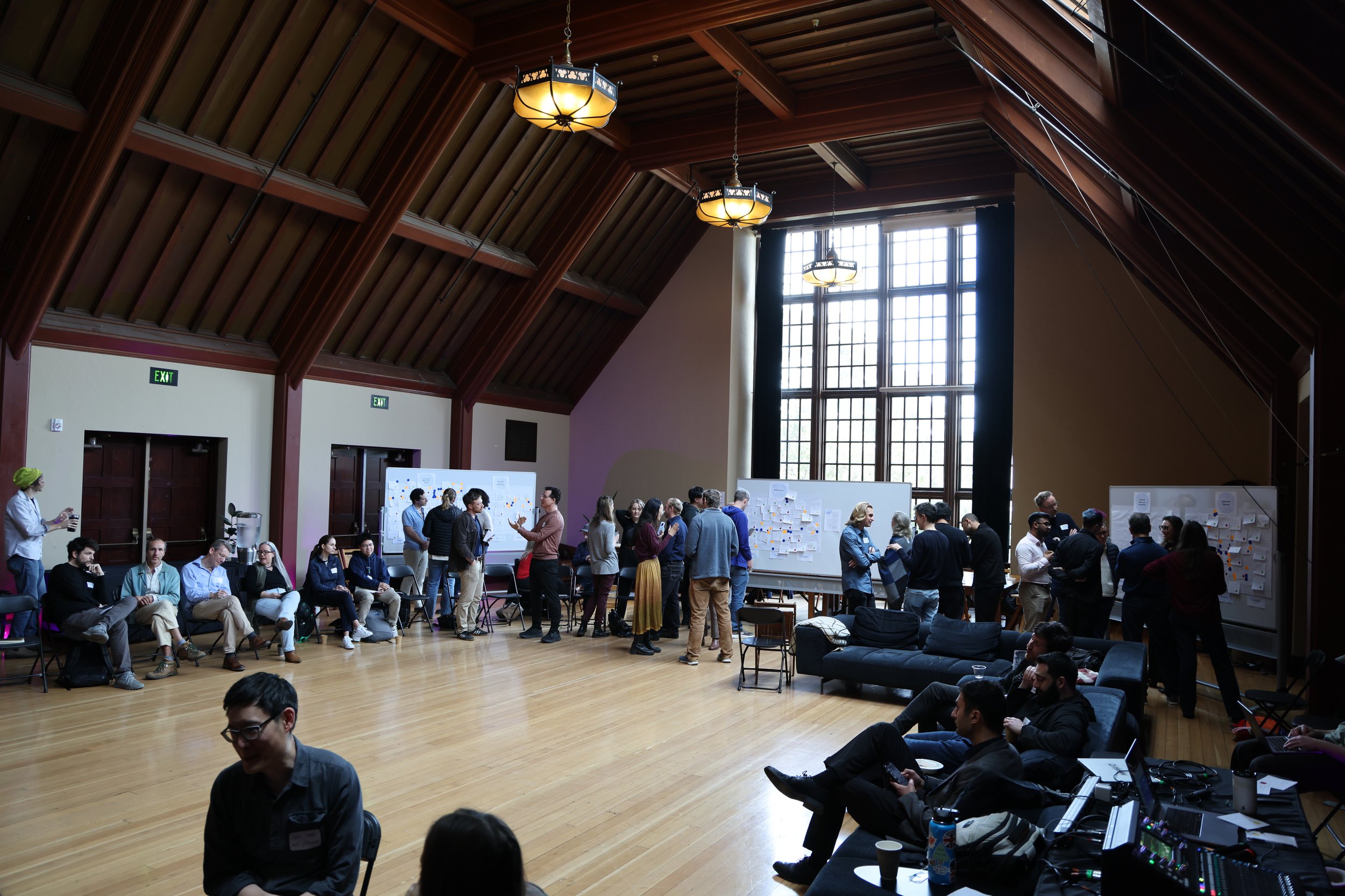 People gathered in a large conference room with high wooden ceiling, large windows, and whiteboards, participating in a workshop or brainstorming session.