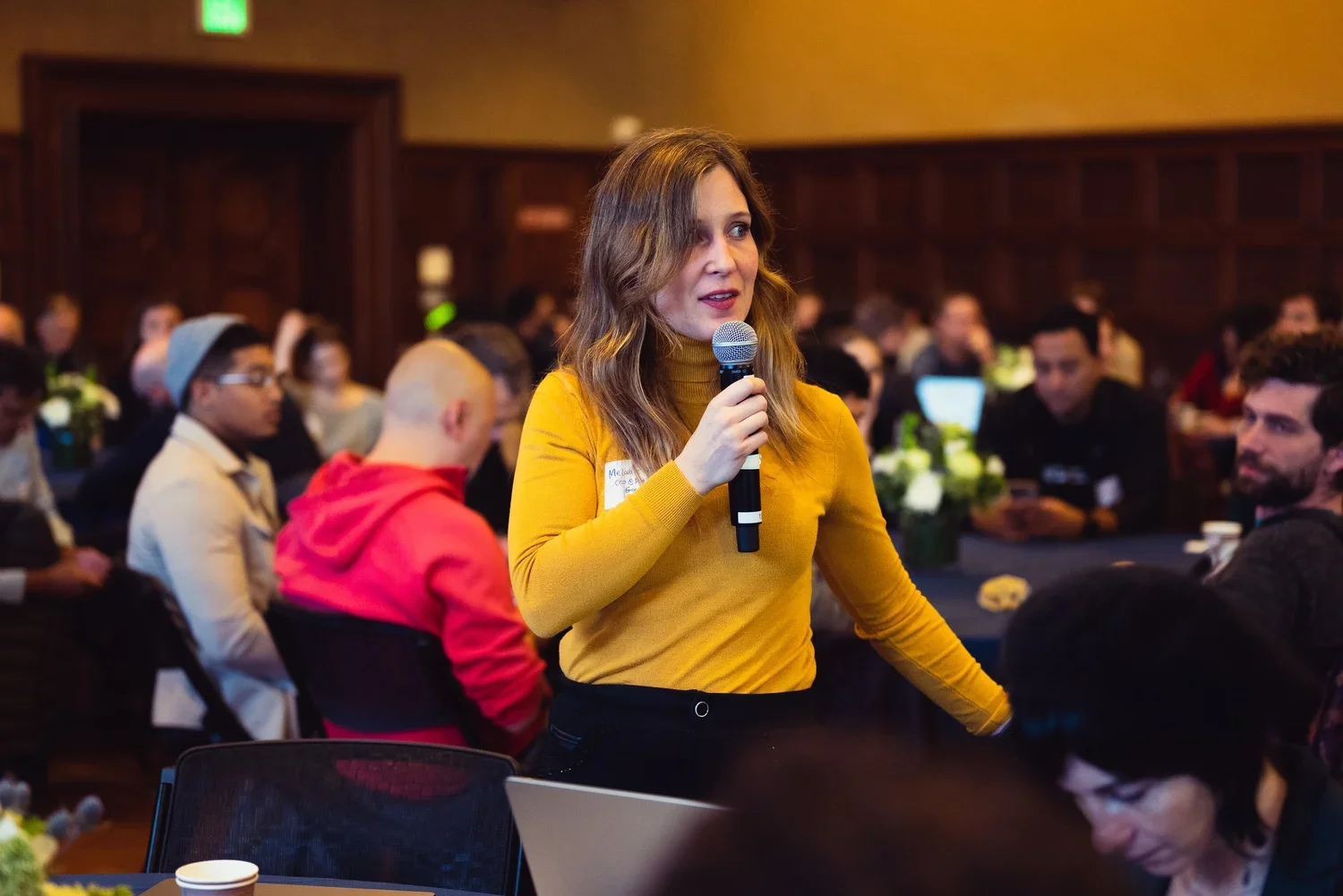 A woman in a yellow sweater speaking into a microphone at a conference or meeting with a diverse group of people seated around tables.