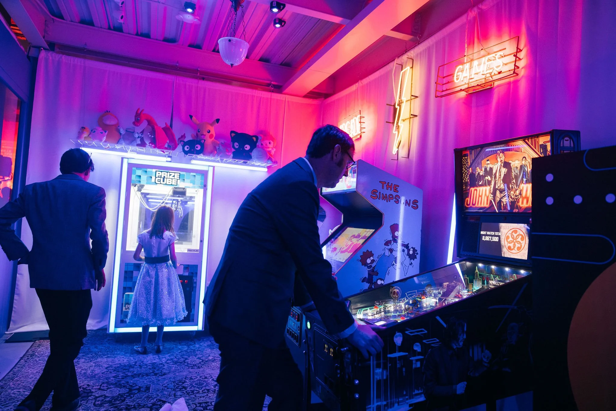 People playing arcade games in a neon-lit arcade room decorated with plush toys and neon signs.