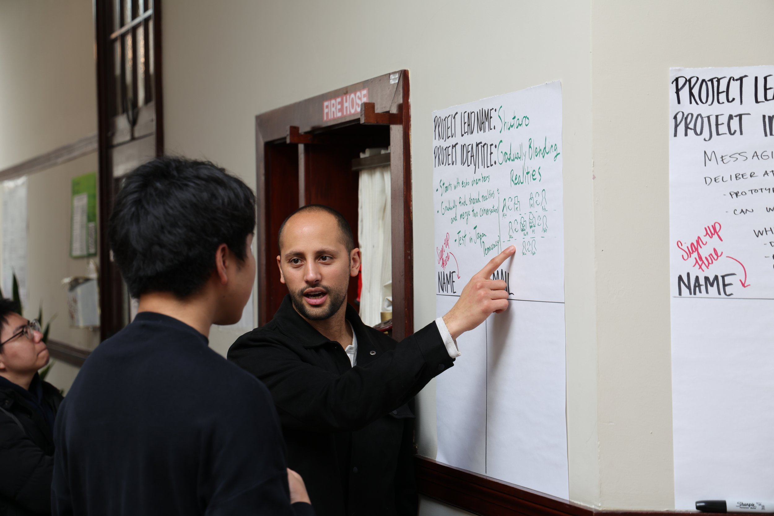 Three people in a discussion, one man pointing at a whiteboard with handwritten notes about a project, while another man listens and a woman observes in the background.
