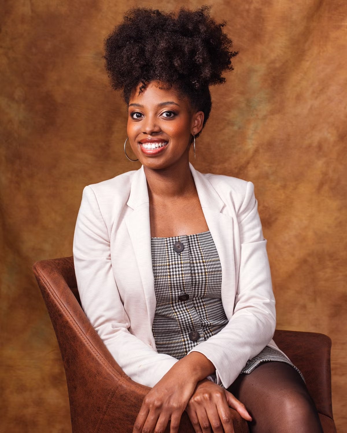 Portrait of a woman with short curly hair, wearing a white blazer over a checkered dress, sitting on a brown chair, smiling against a brownish background.