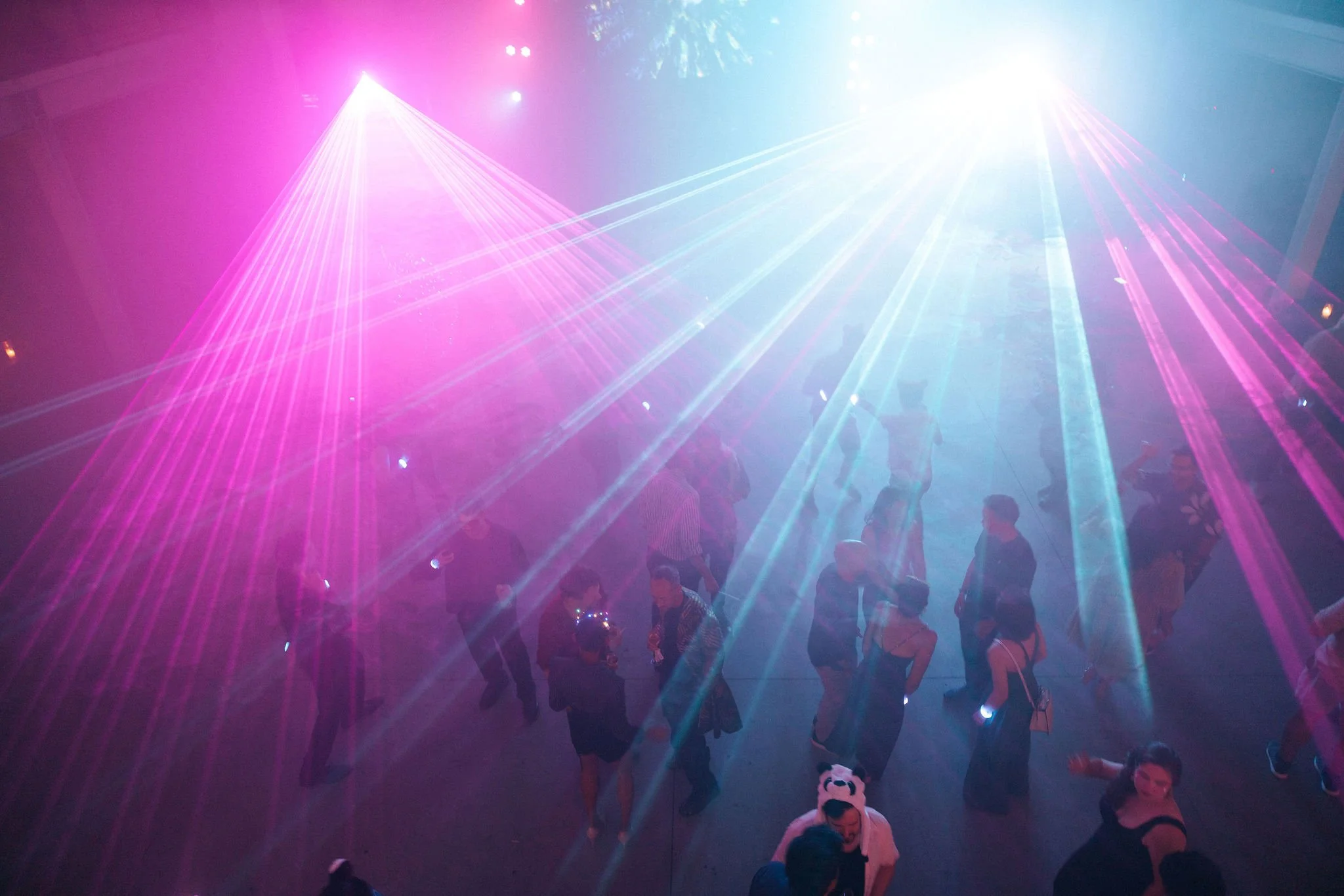 People dancing and socializing on a dance floor with pink and blue laser lights illuminating the scene.